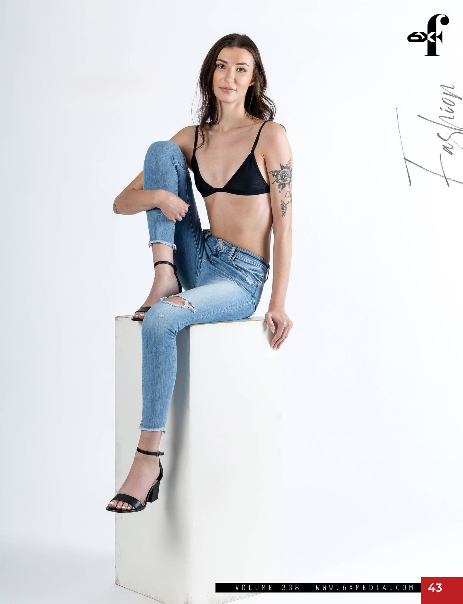 Brunette model sits on white cube wearing black triangle bralette and ripped blue jeans with heels.