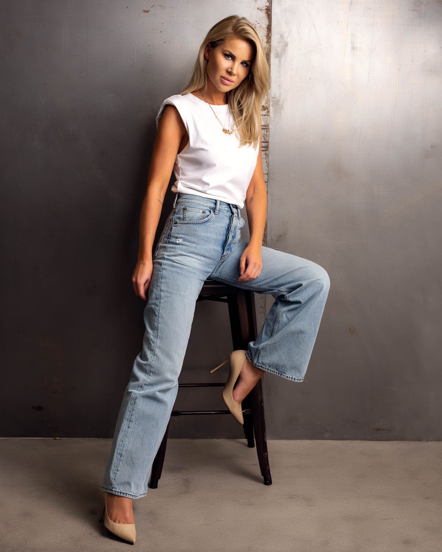 Blonde woman in white top and light wash jeans poses on stool in studio.