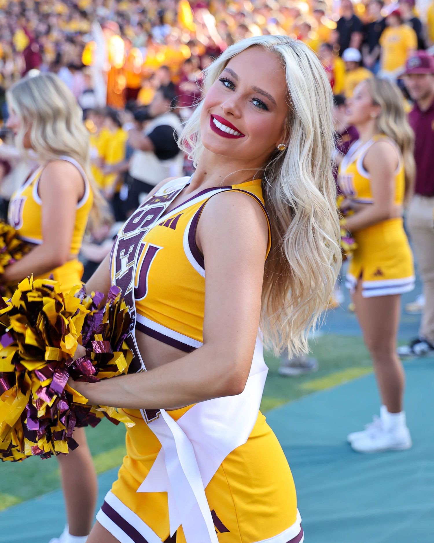 Smiling blonde cheerleader in gold uniform performing at stadium event.