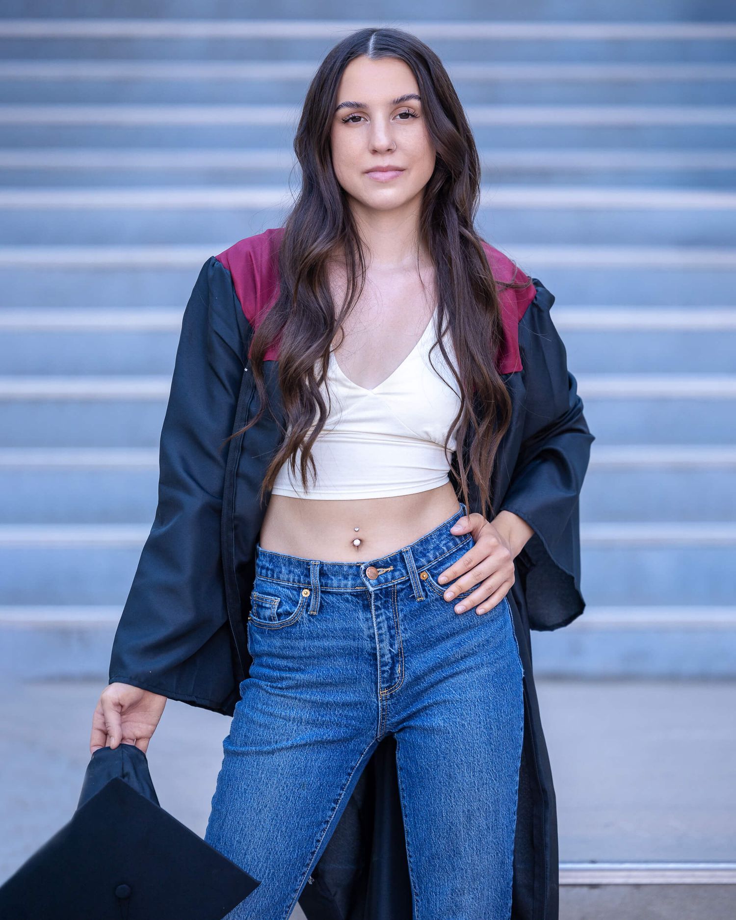 Young woman in graduation gown, white crop top, and jeans holding a cap on steps.