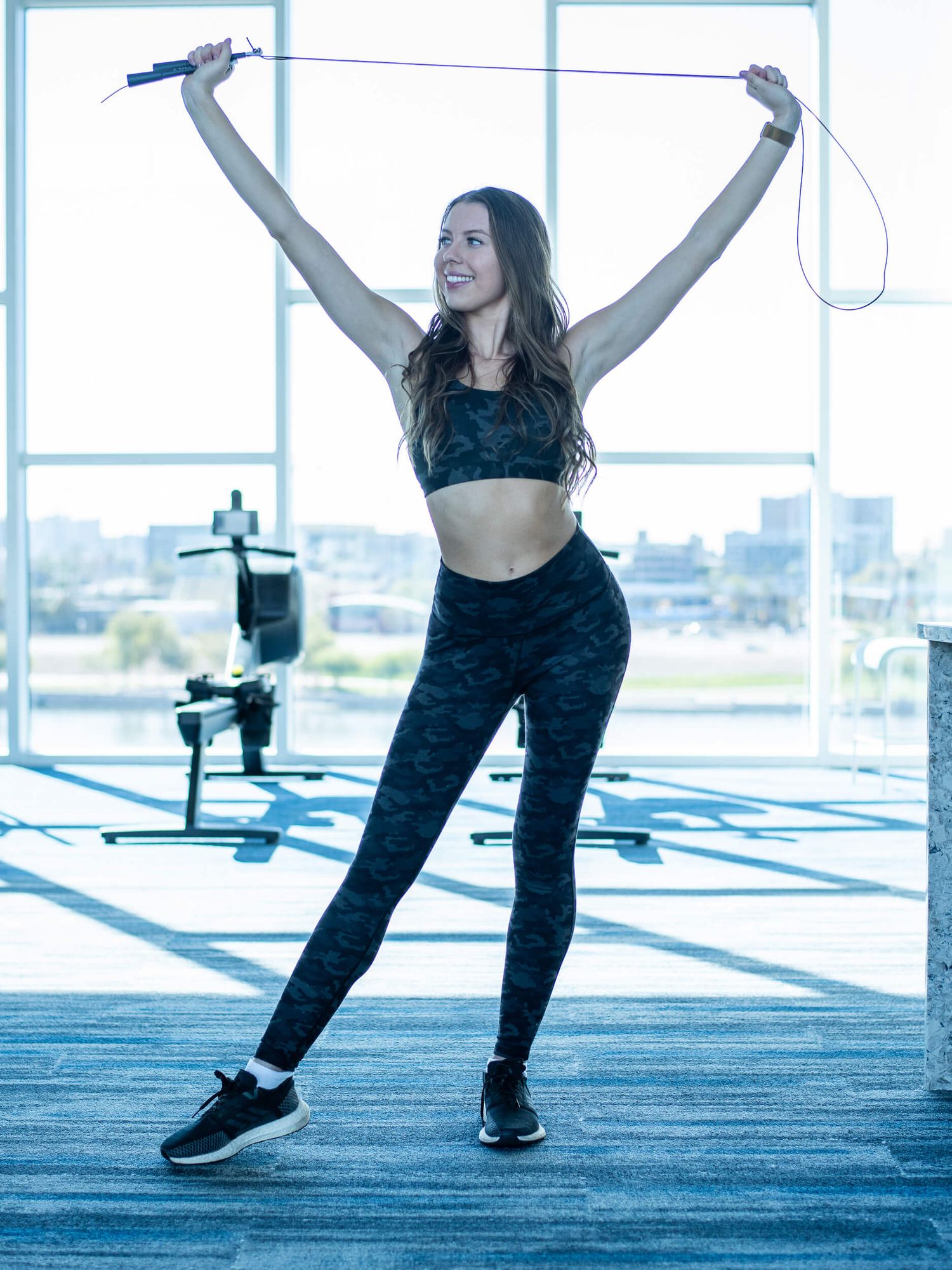 Woman in camo leggings and sports bra celebrates with jump rope raised overhead in bright modern gym.
