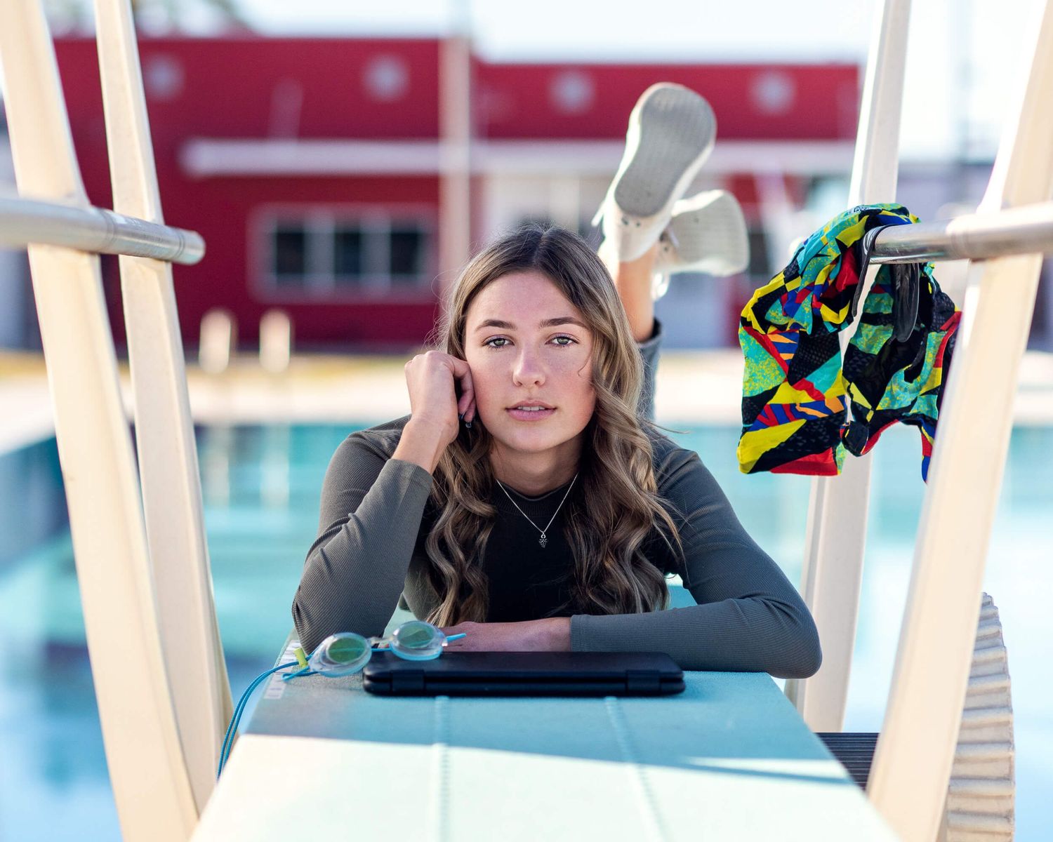 Woman with wavy hair relaxes poolside on a diving board with a laptop, goggles, and colorful swimsuit nearby.