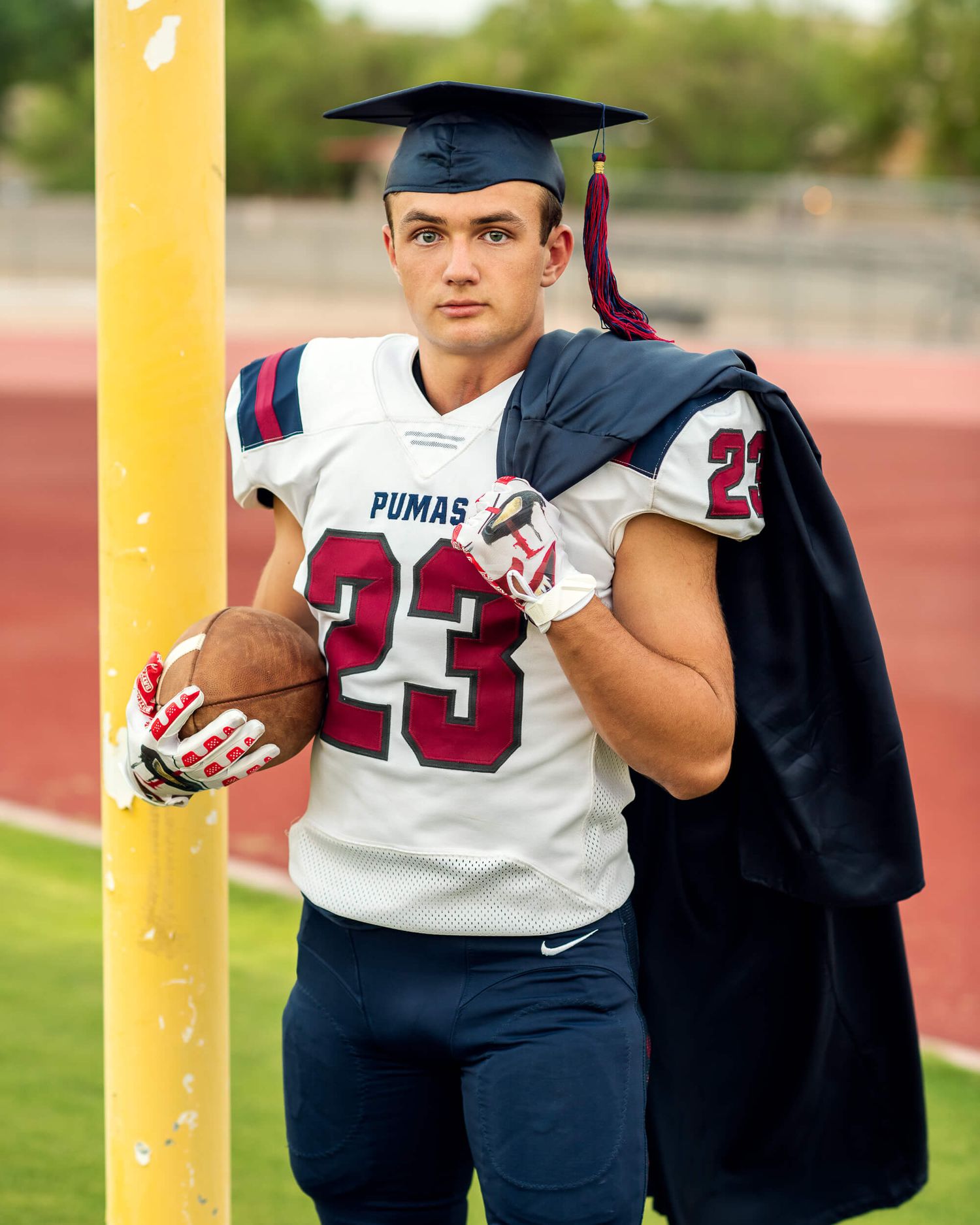 High school senior in Pumas football jersey number 23 holding ball wearing graduation cap and gown at stadium.