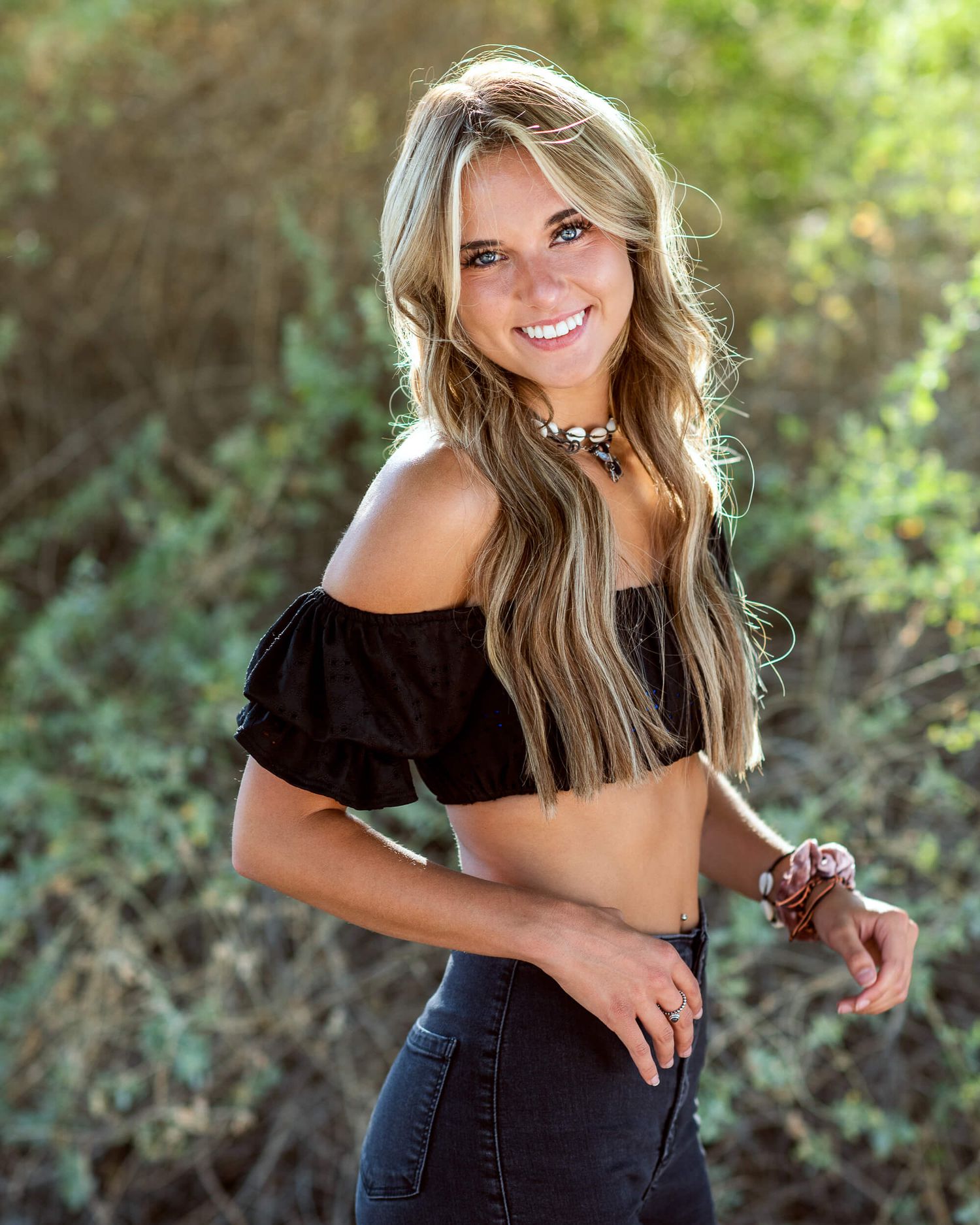 Smiling young woman with long blonde hair wearing a black off-shoulder crop top and jeans, posing outdoors.