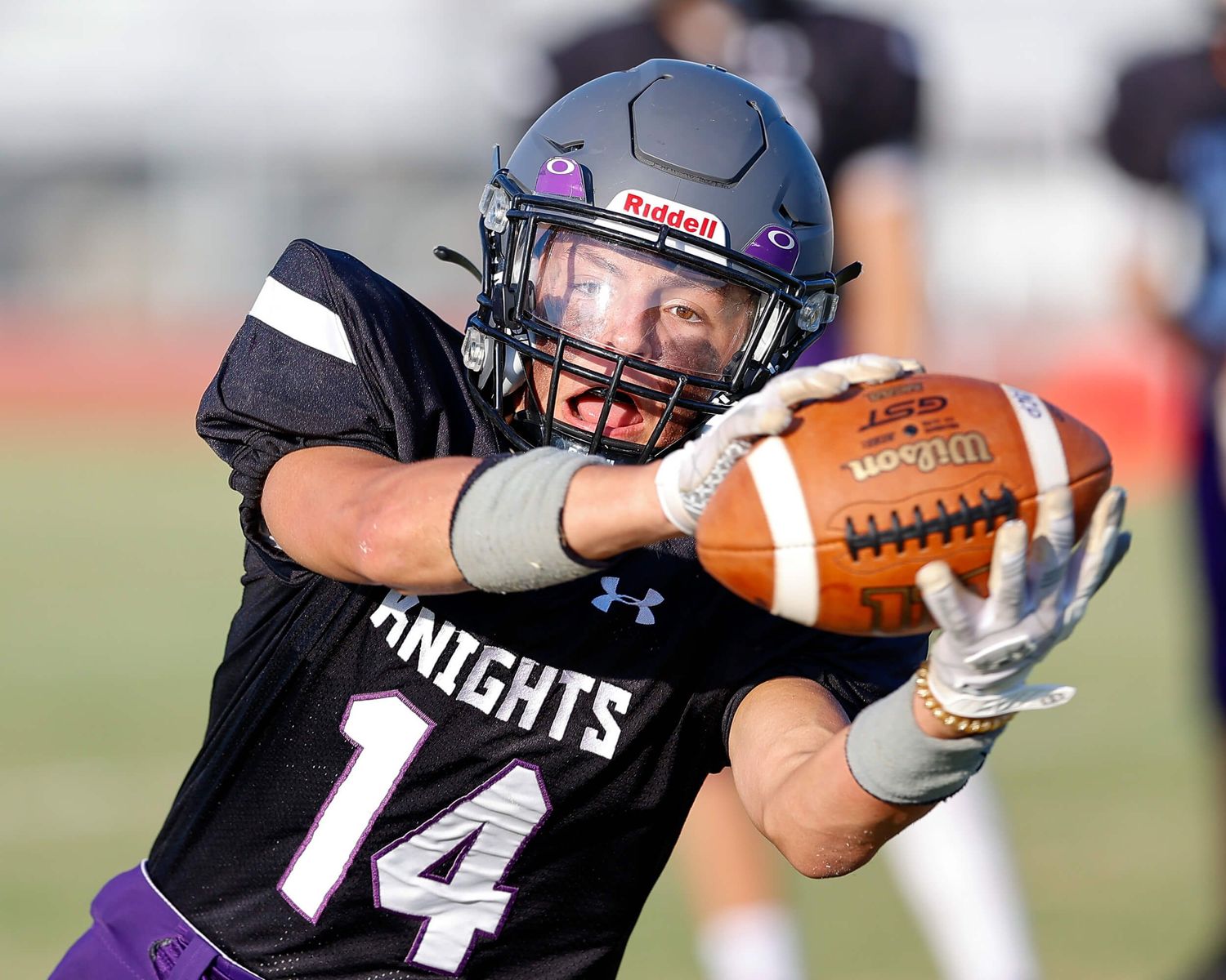 Football player wearing Knights jersey number 14 catches a pass during a high school game under bright sunlight.