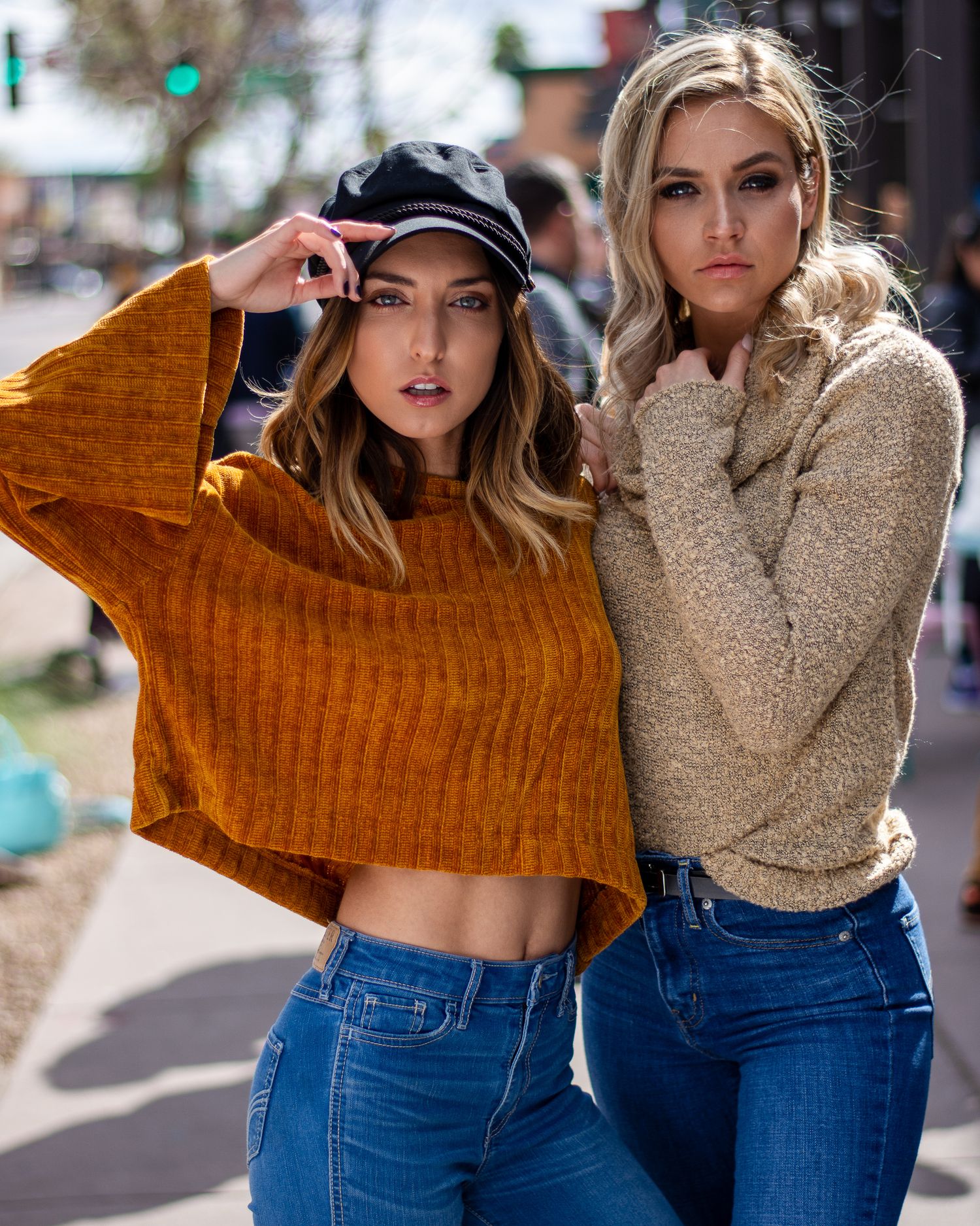 Two young women in knit sweaters and jeans pose together on a city street in casual fall fashion.