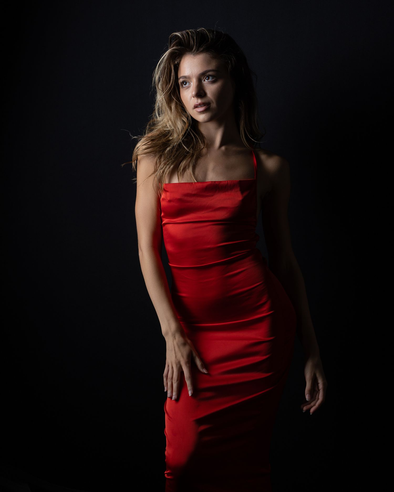 Young woman poses in elegant red satin fitted dress against dark studio background.