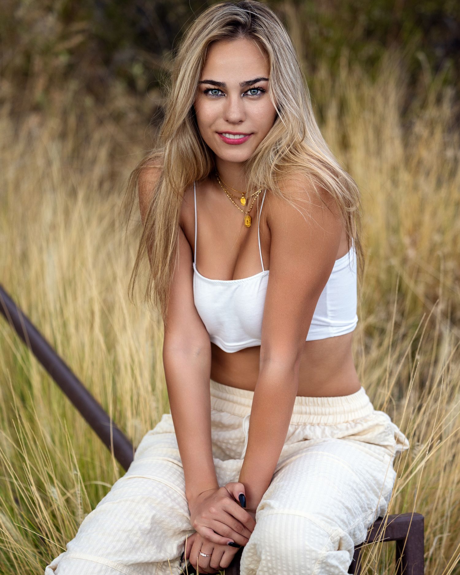 Blonde woman with blue eyes wearing white crop top and linen pants sitting outdoors in tall grass.