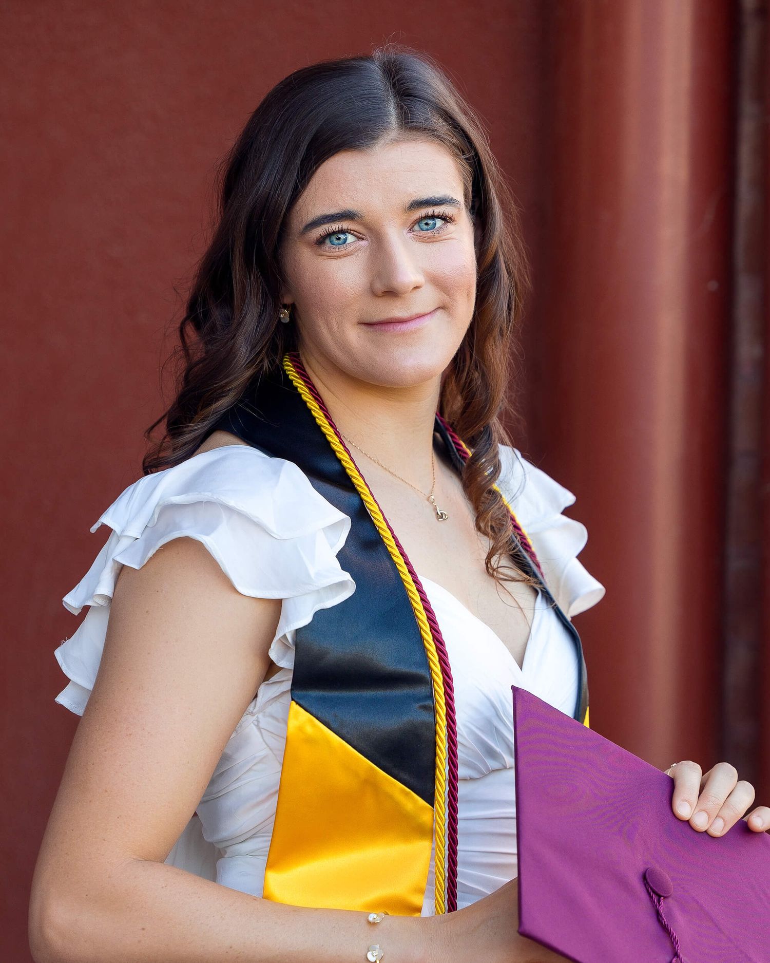 Young woman in white ruffled dress wearing colorful graduation stole holding maroon cap near brick wall.