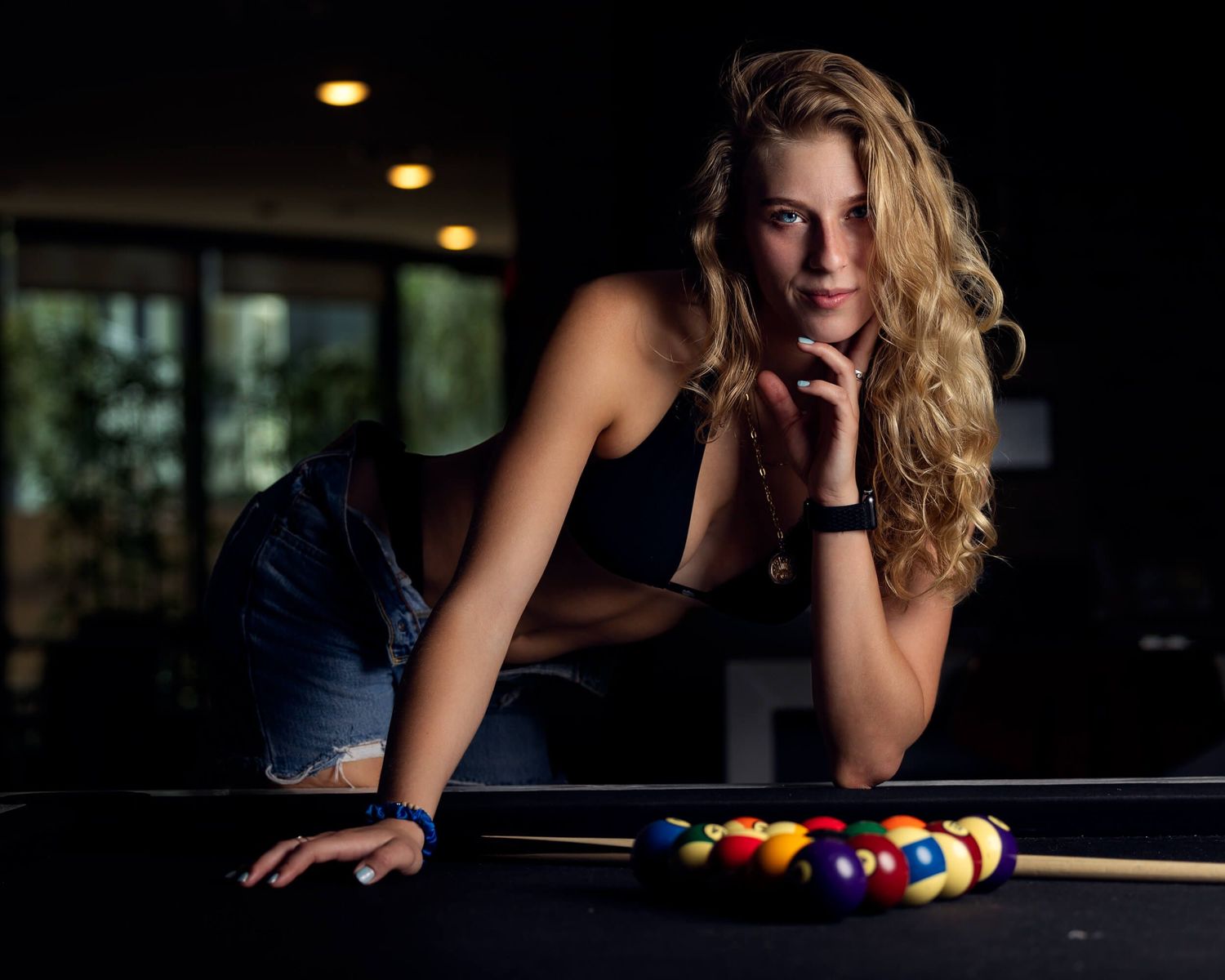 Curly blonde woman in denim leans over pool table with billiard balls in dim light.