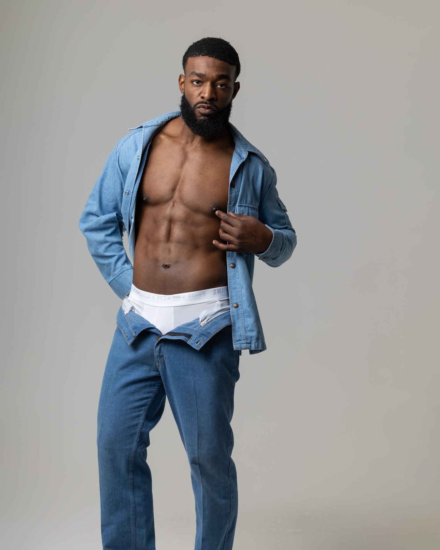 Muscular Black man with beard posing shirtless in open denim shirt and jeans, showing toned abs.