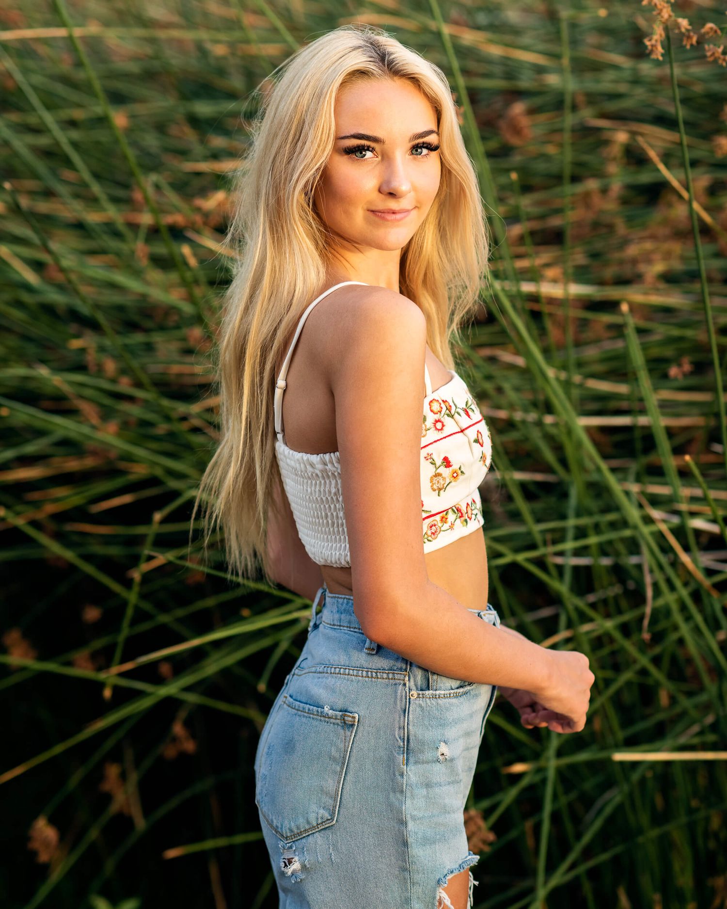 Blonde woman in ripped jeans and a crop top posing in a grassy field at golden hour.