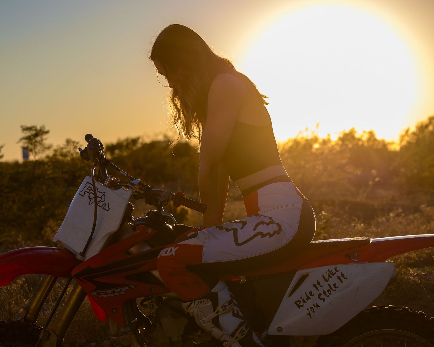 Woman sitting on a dirt bike silhouetted against a golden sunset in an outdoor setting.