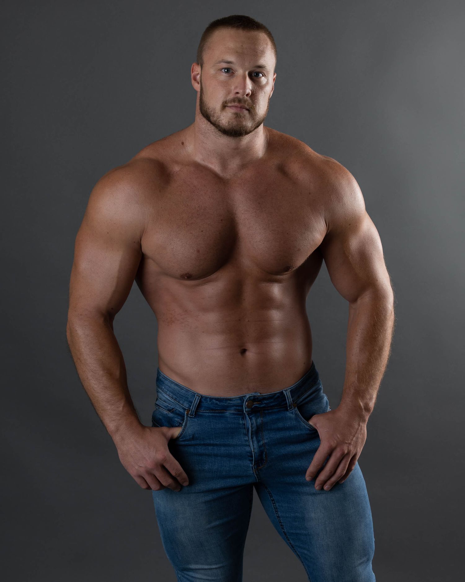Muscular shirtless male bodybuilder posing in blue jeans on gray background.