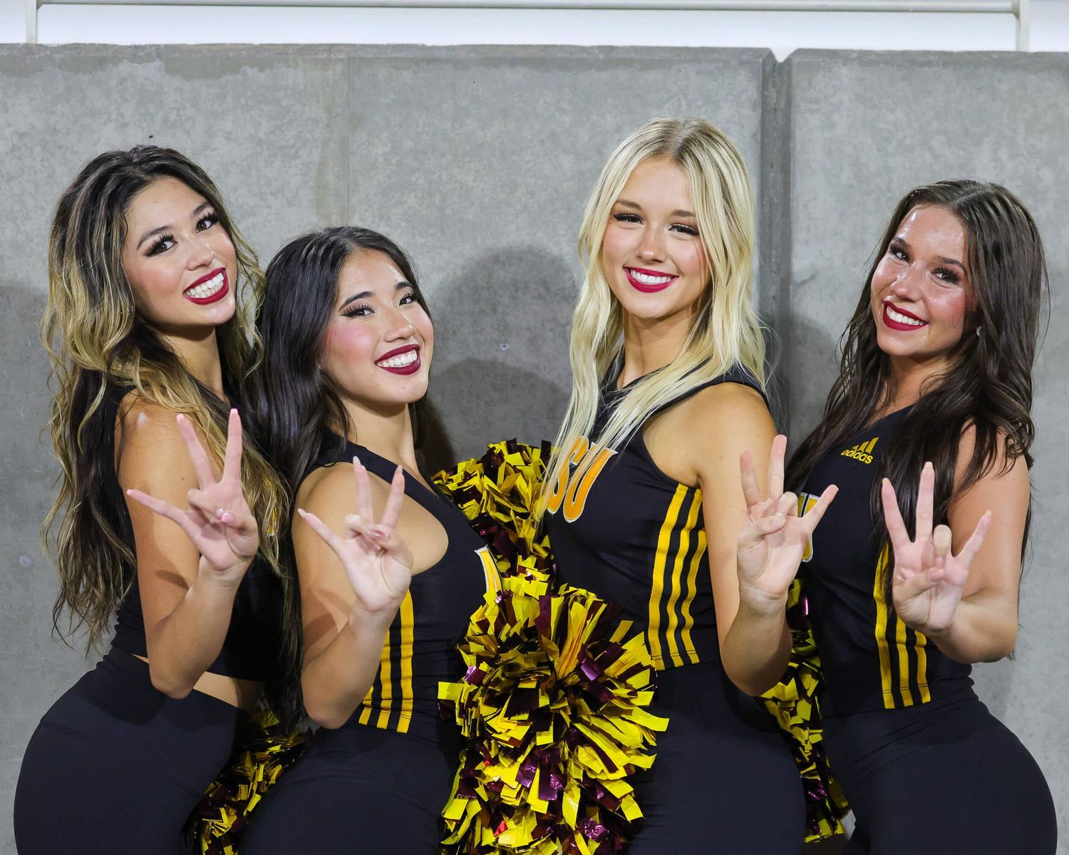 Four ASU cheerleaders in black uniforms pose together holding gold pom-poms.