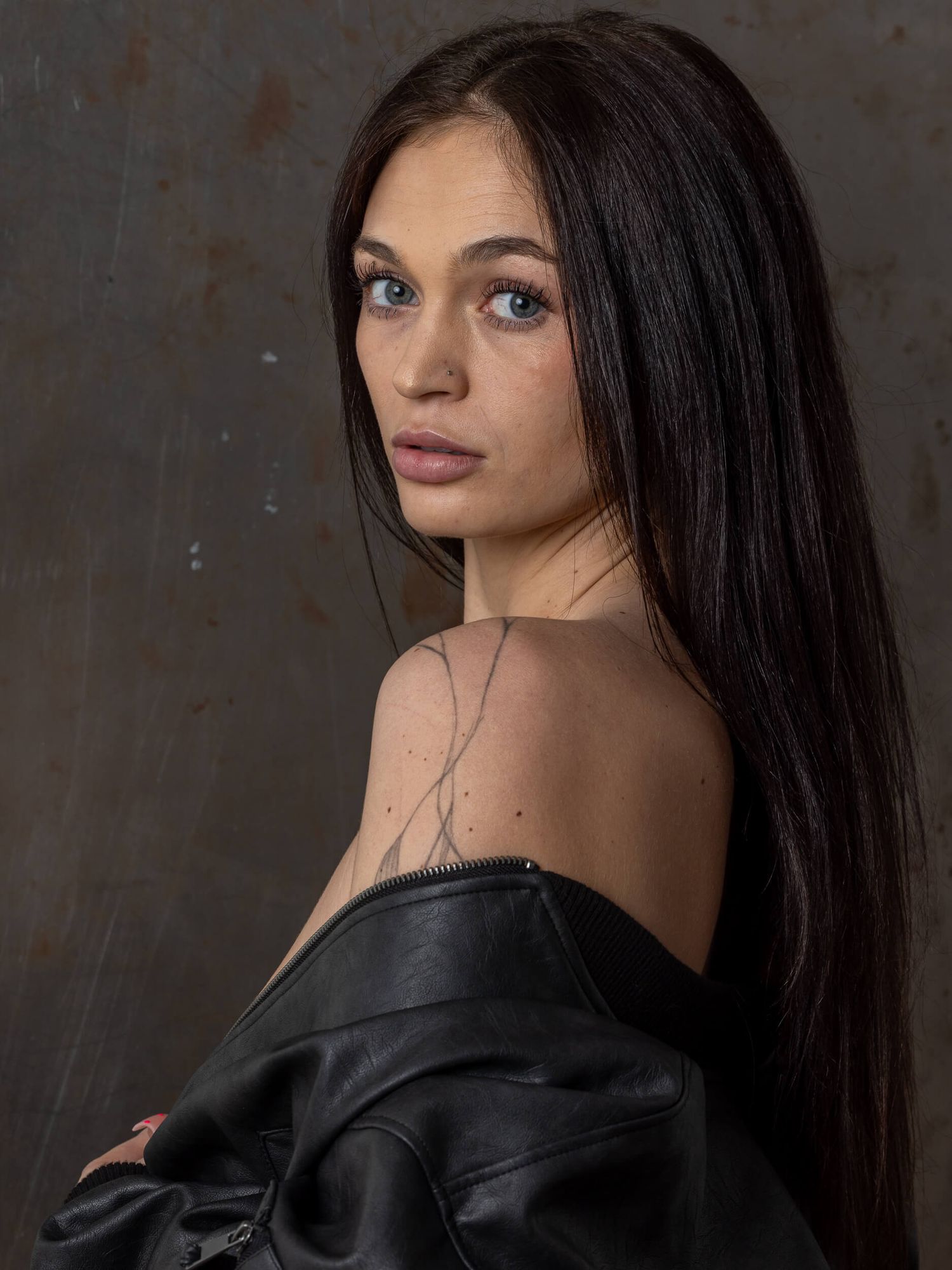 Young woman with long dark hair in black leather off-shoulder jacket poses against dark textured background.