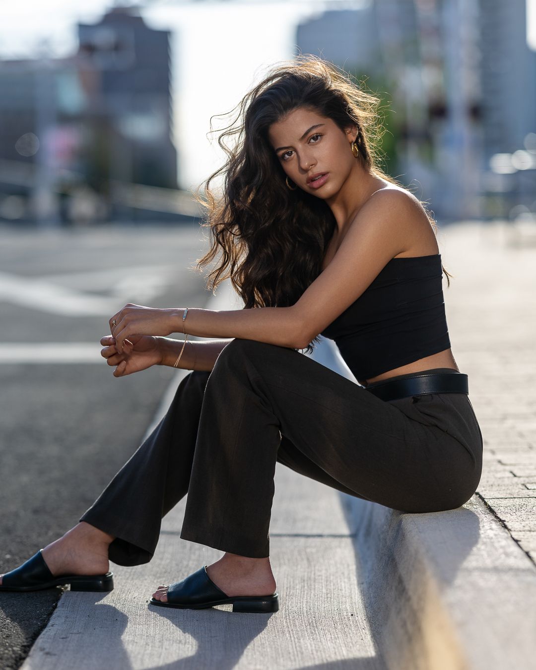 Brunette woman in black crop top and pants sitting on urban curb in city setting.