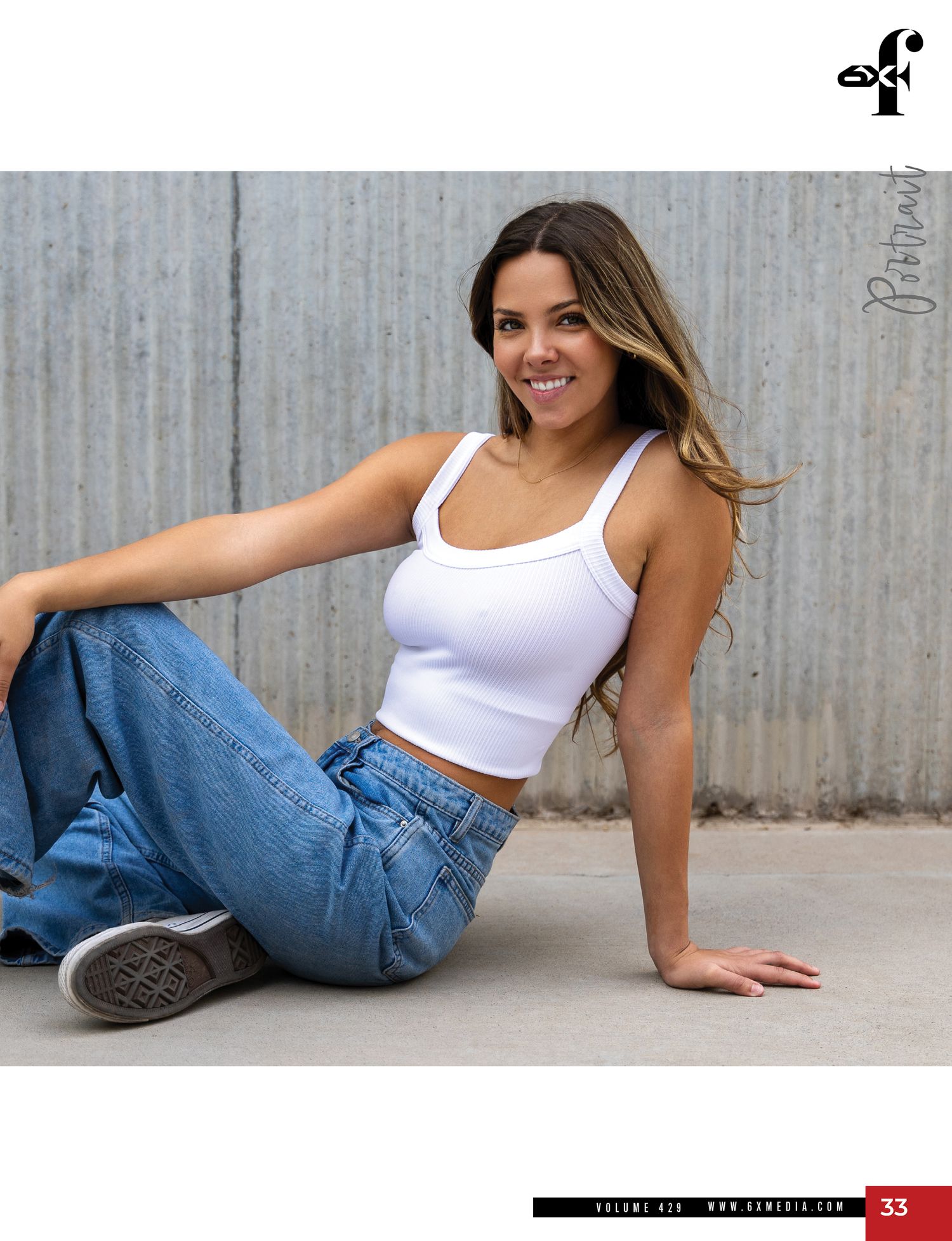 Smiling young woman in white crop top and jeans sits casually on the ground for a magazine shoot.