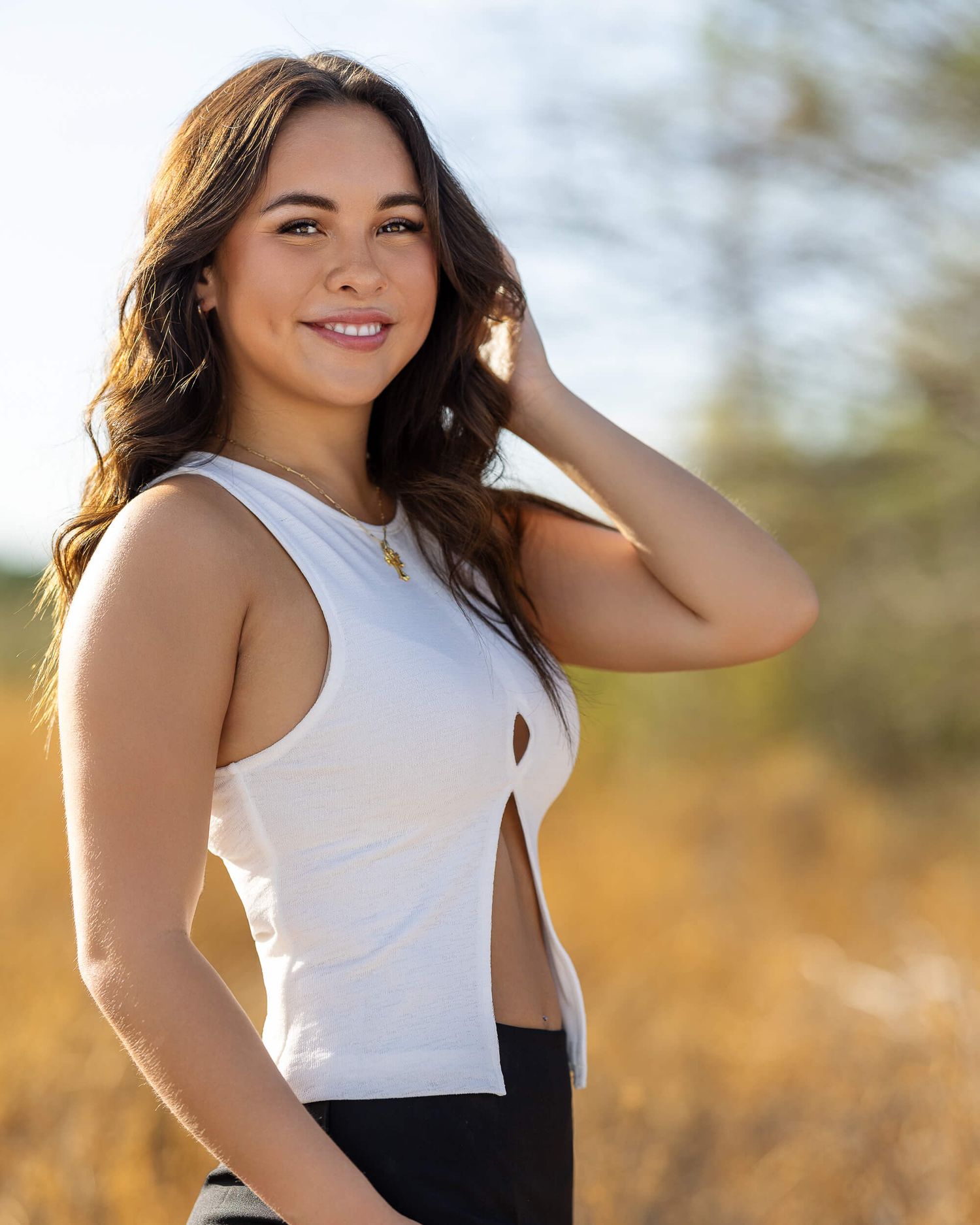 Young brunette woman smiling in white crop top posing outdoors in golden hour sunlight.