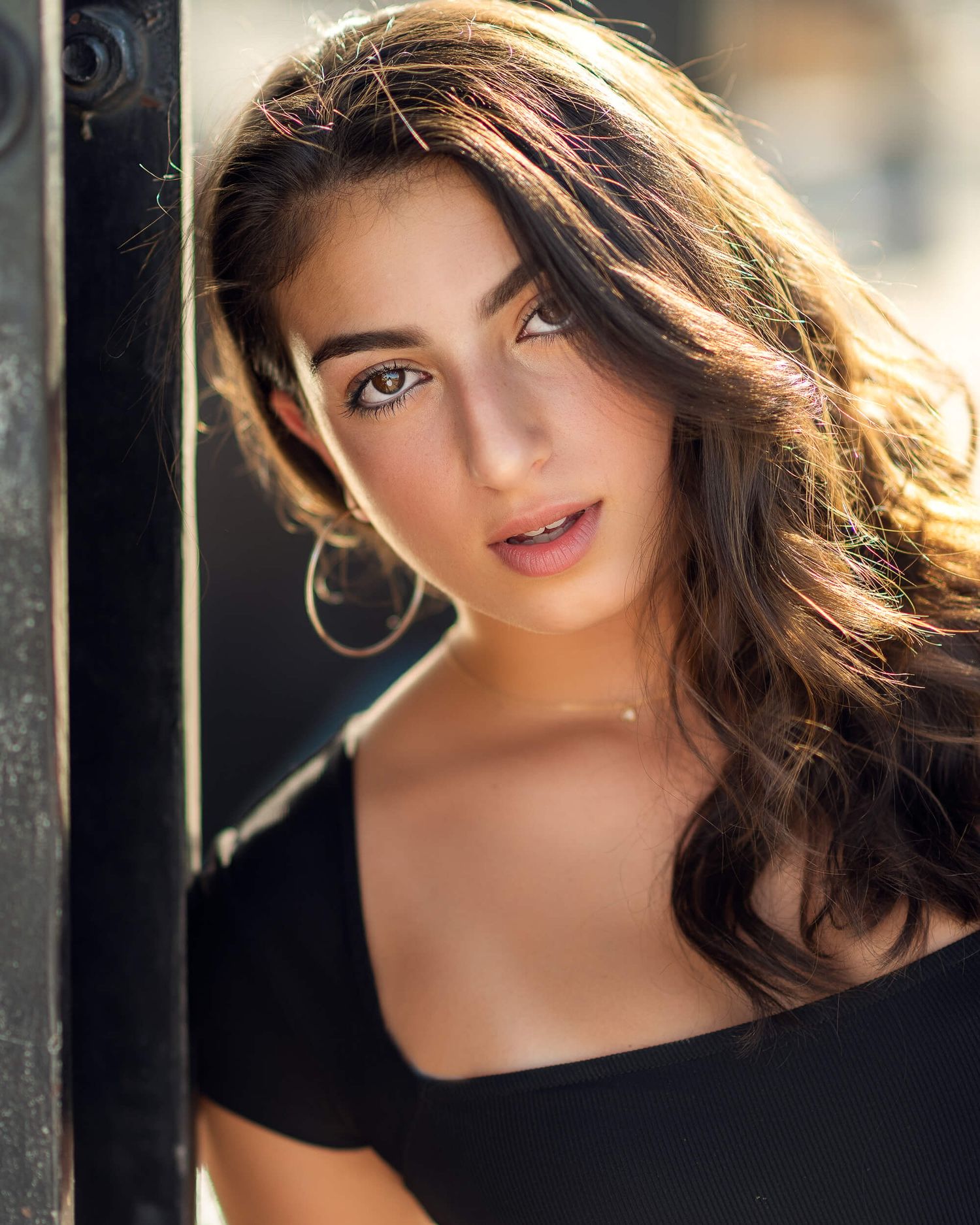 Young woman with long dark wavy hair wearing a black off-shoulder top in warm outdoor lighting.