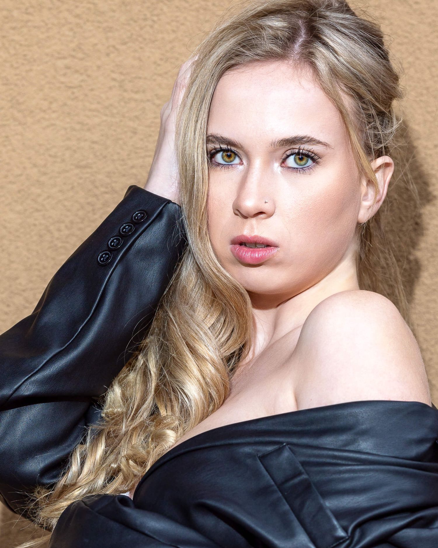 Blonde woman with green eyes wearing off-shoulder black blazer posing against beige background.