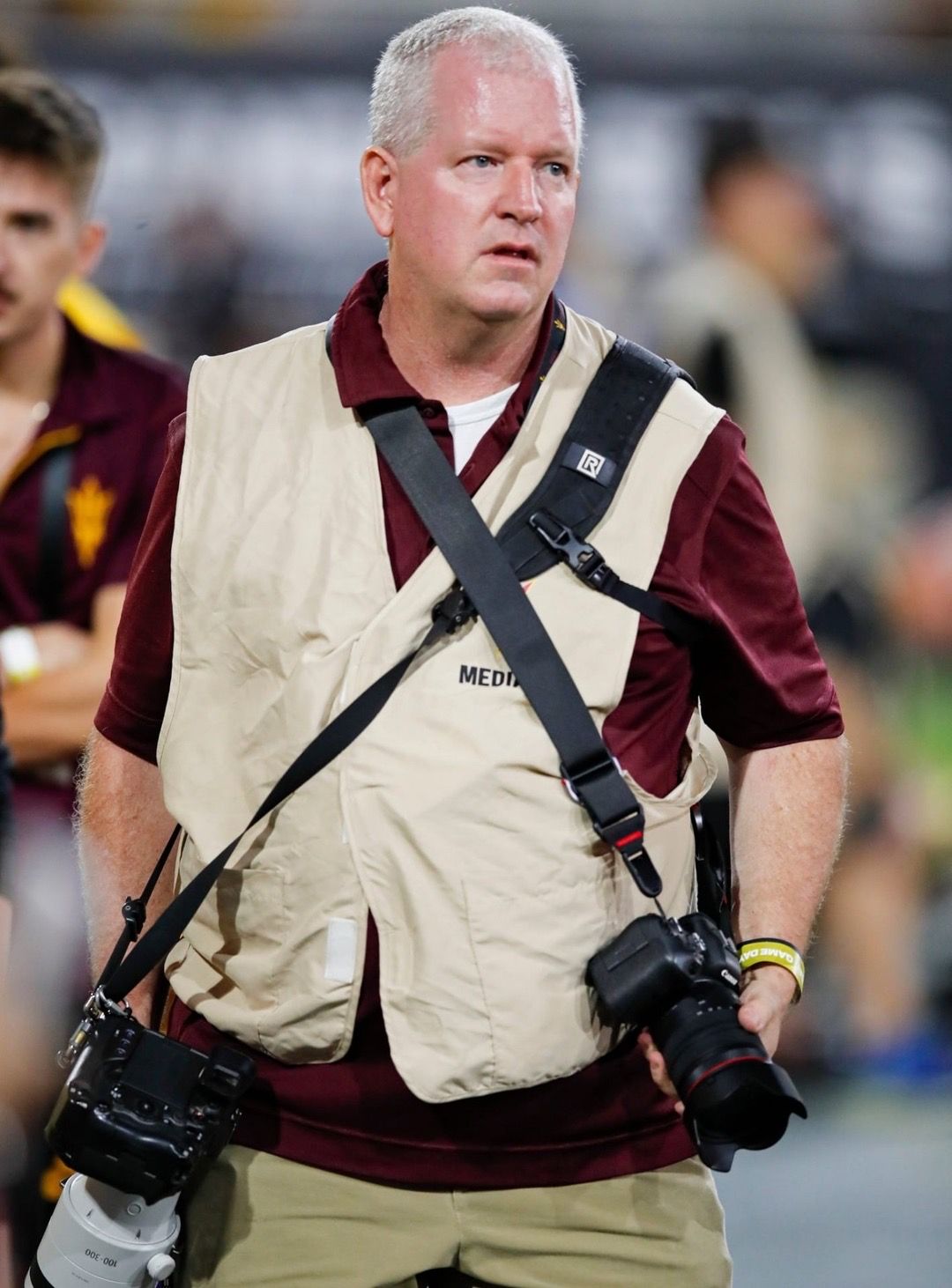 A media photographer wearing a vest and carrying two cameras at a sports event.