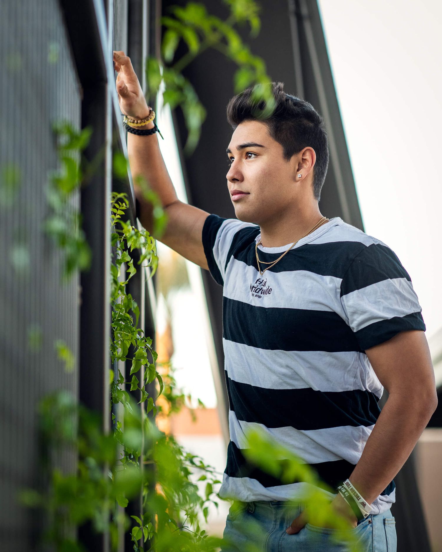 Young man in striped black and white t-shirt leaning on railing surrounded by green plants outdoors.