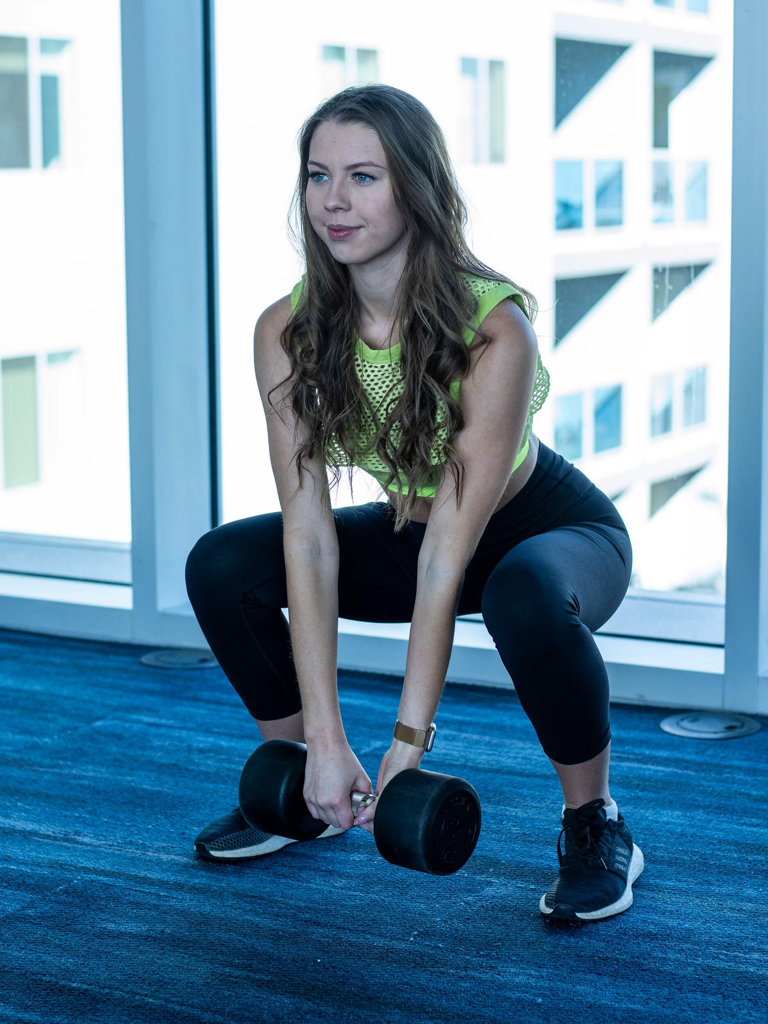 Woman in green top and black leggings performing a squat with dumbbells in a modern gym with large windows.