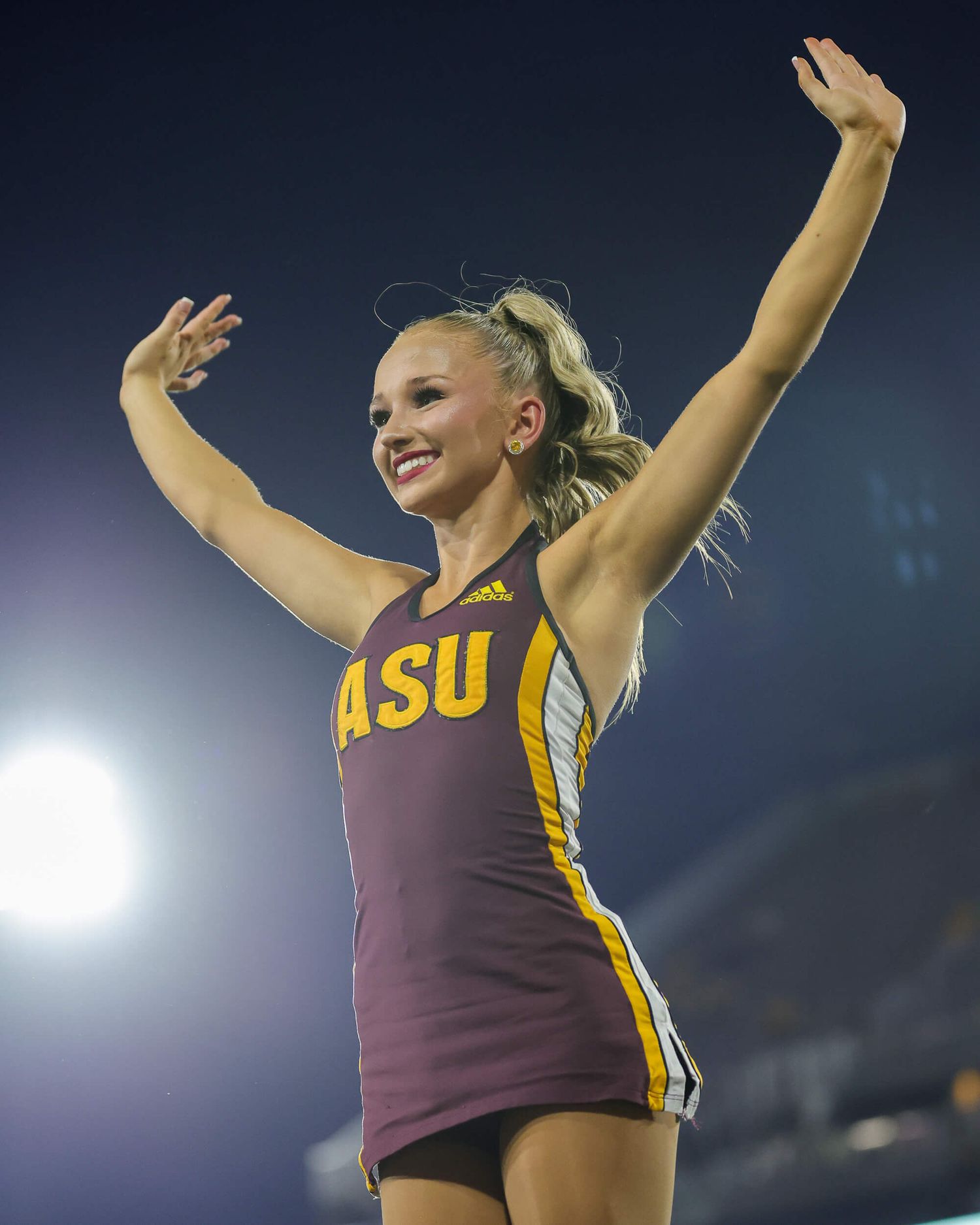 ASU cheerleader in maroon Adidas uniform raises arms enthusiastically during an Arizona State University football game.