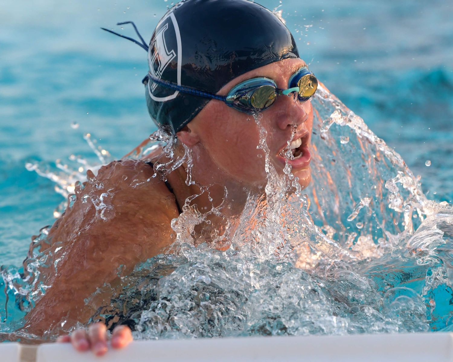Competitive swimmer wearing black cap and goggles emerges from the water during an outdoor swimming race.