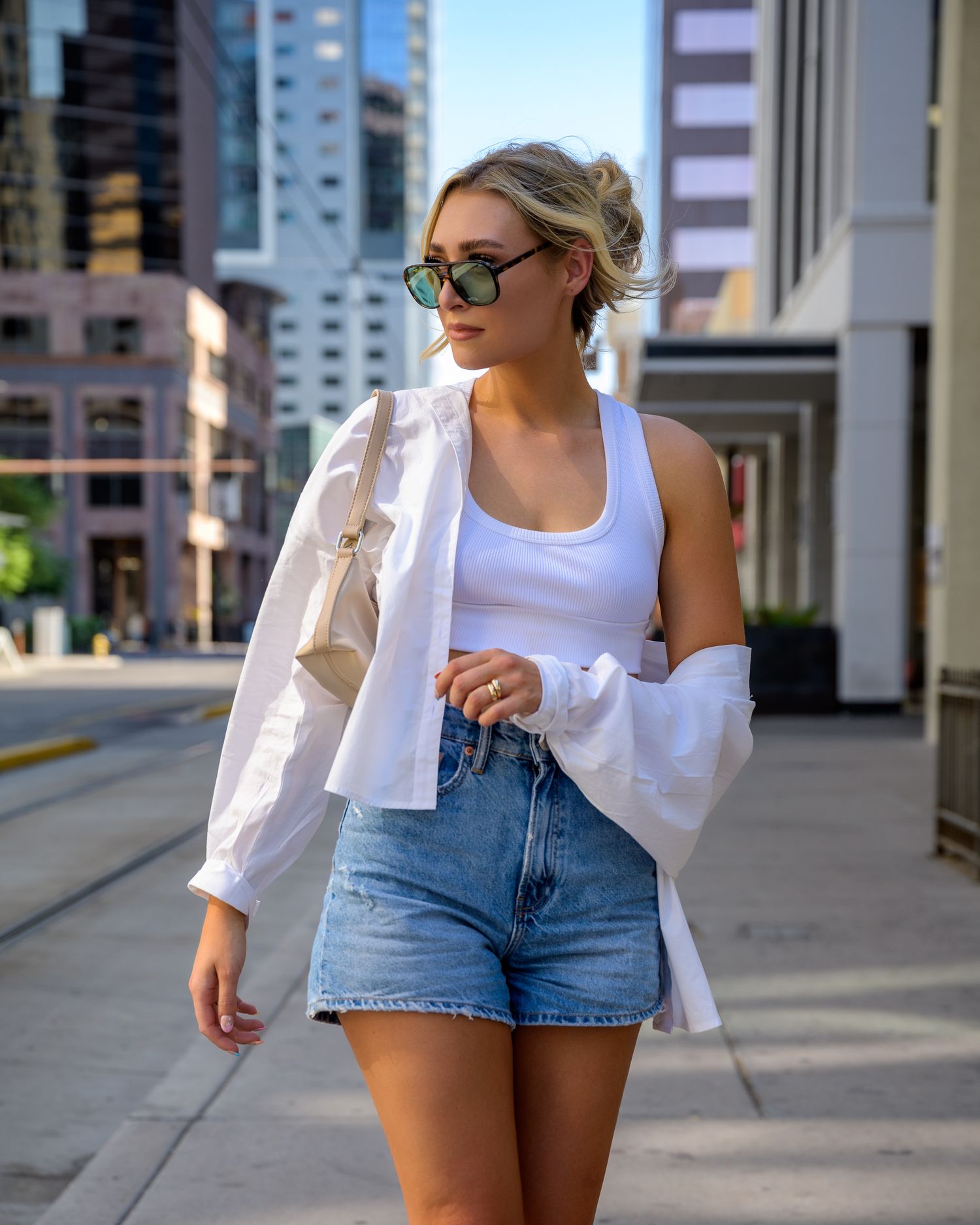 Stylish blonde woman in white top and denim shorts walking on a sunny urban street.
