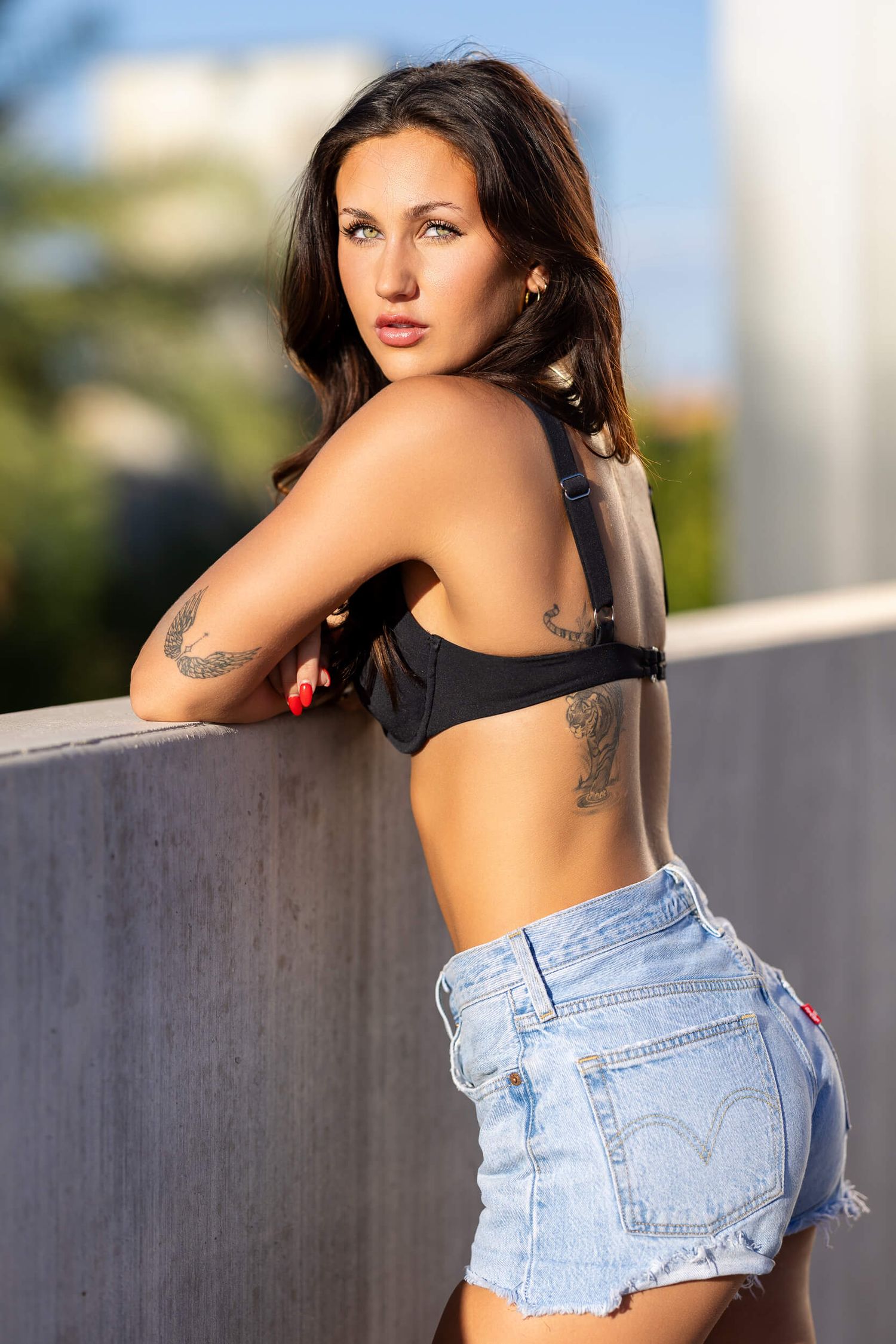 Brunette woman with tattoos wearing black crop top and denim shorts leaning on concrete wall outdoors.