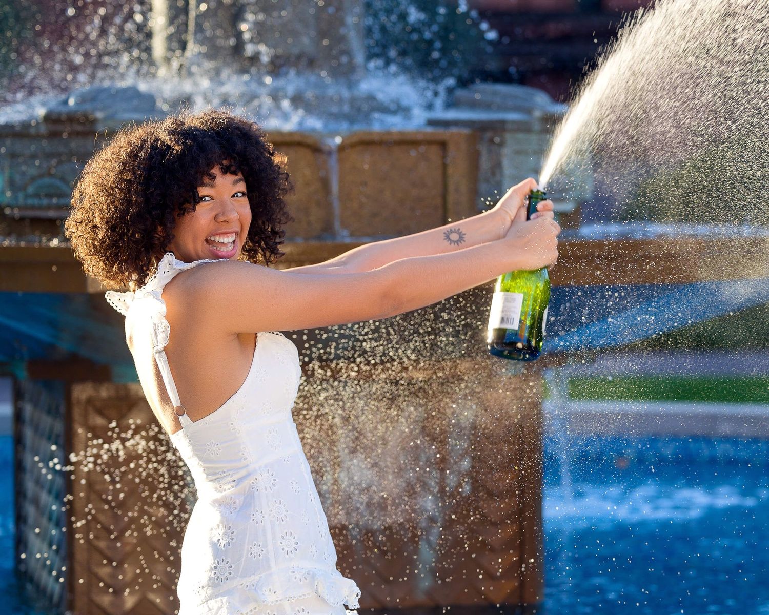 A graduate in white dress celebrates by spraying champagne near a decorative fountain outdoors.