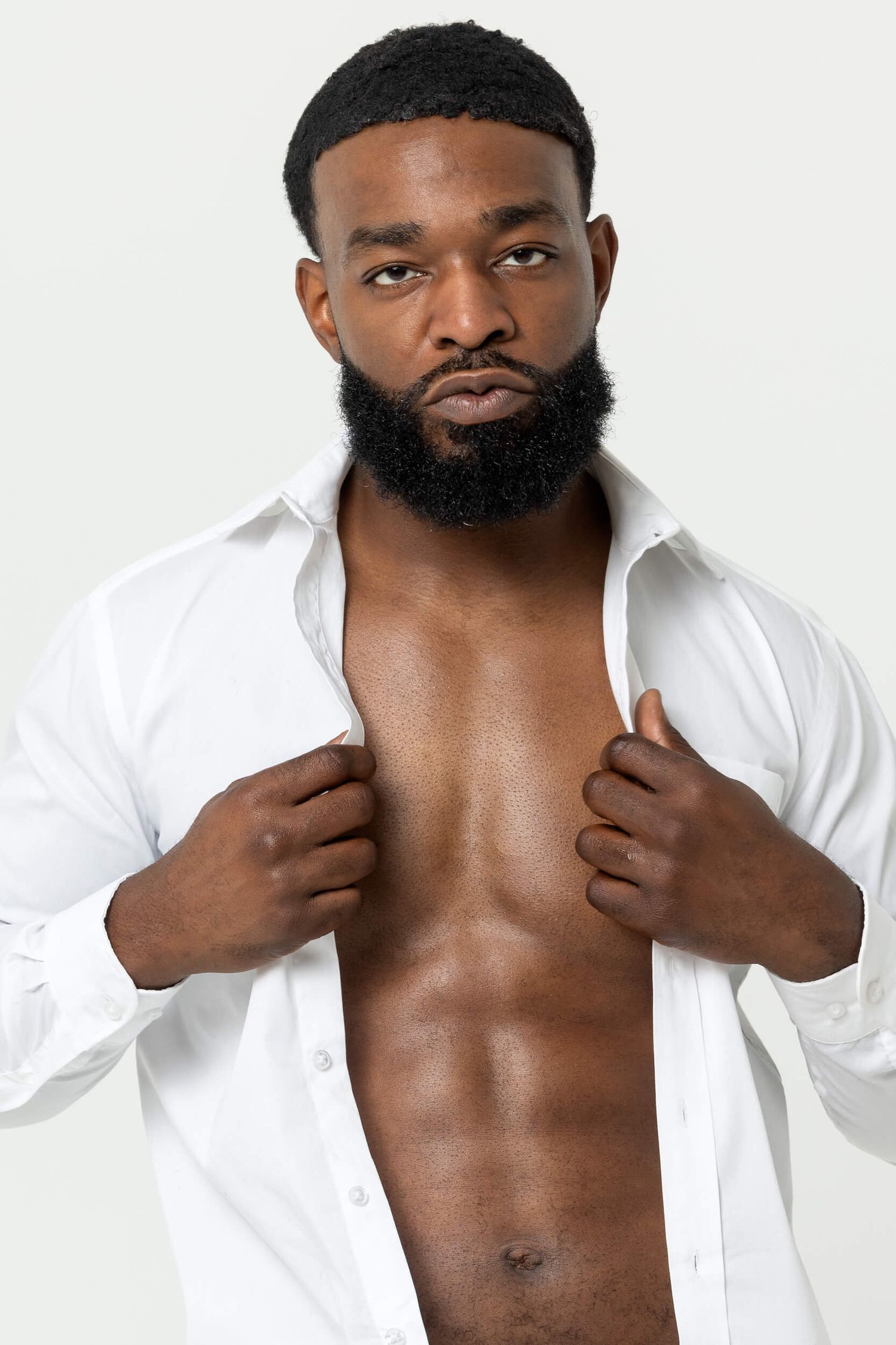 Muscular Black man with beard opening white dress shirt revealing toned physique against light gray background.