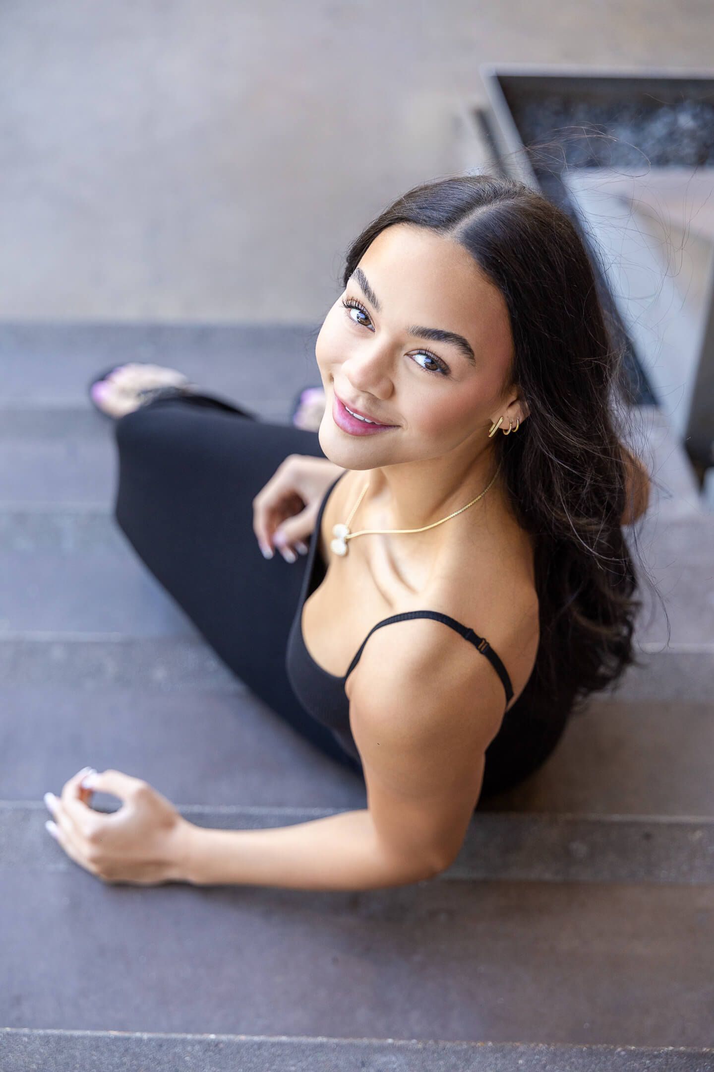 Young woman in black outfit sitting on steps wearing gold jewelry and smiling at camera.