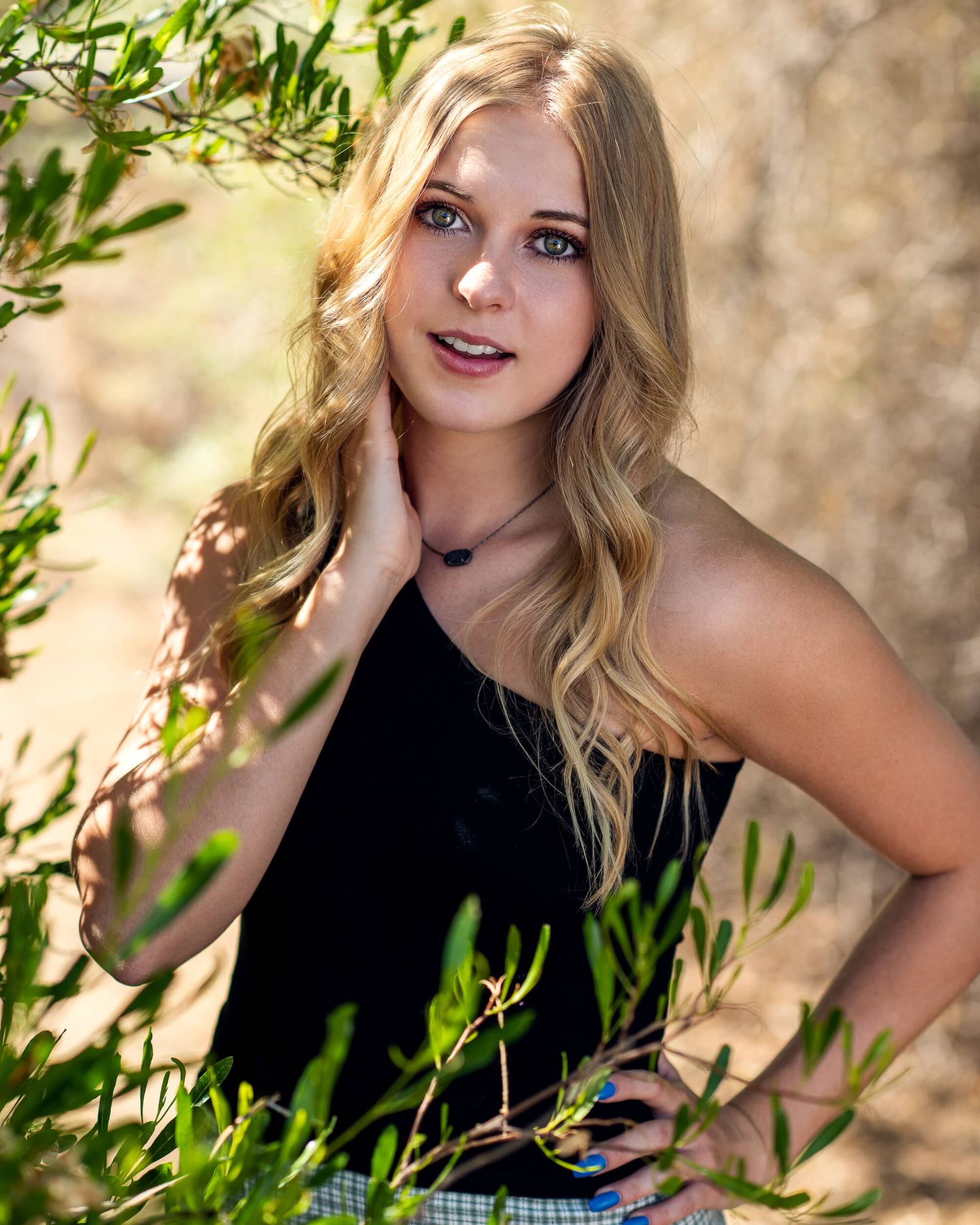 Young blonde woman in black top poses among green branches outdoors in warm sunlight.