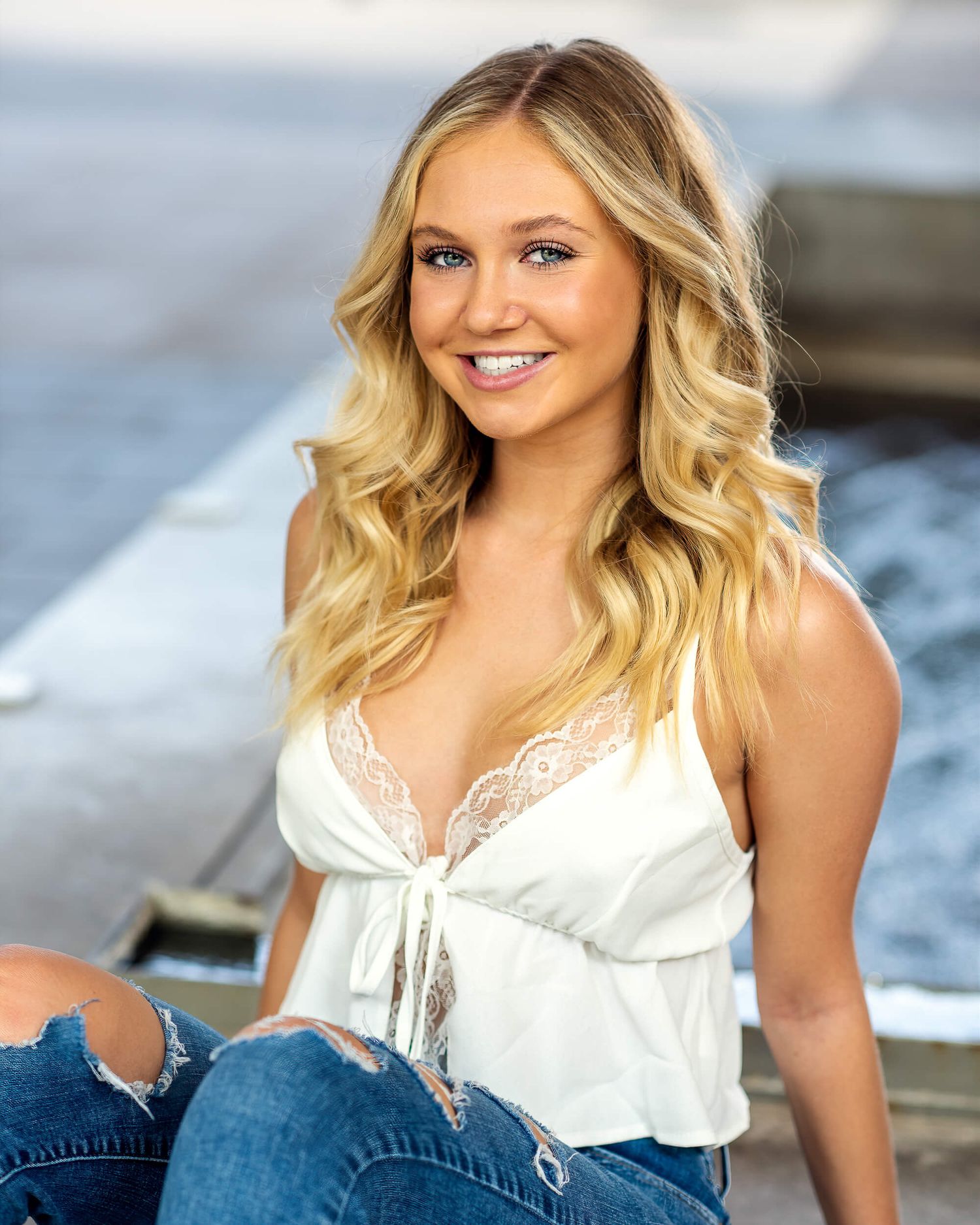 Smiling blonde woman in a white lace top and ripped jeans sitting outdoors near water.