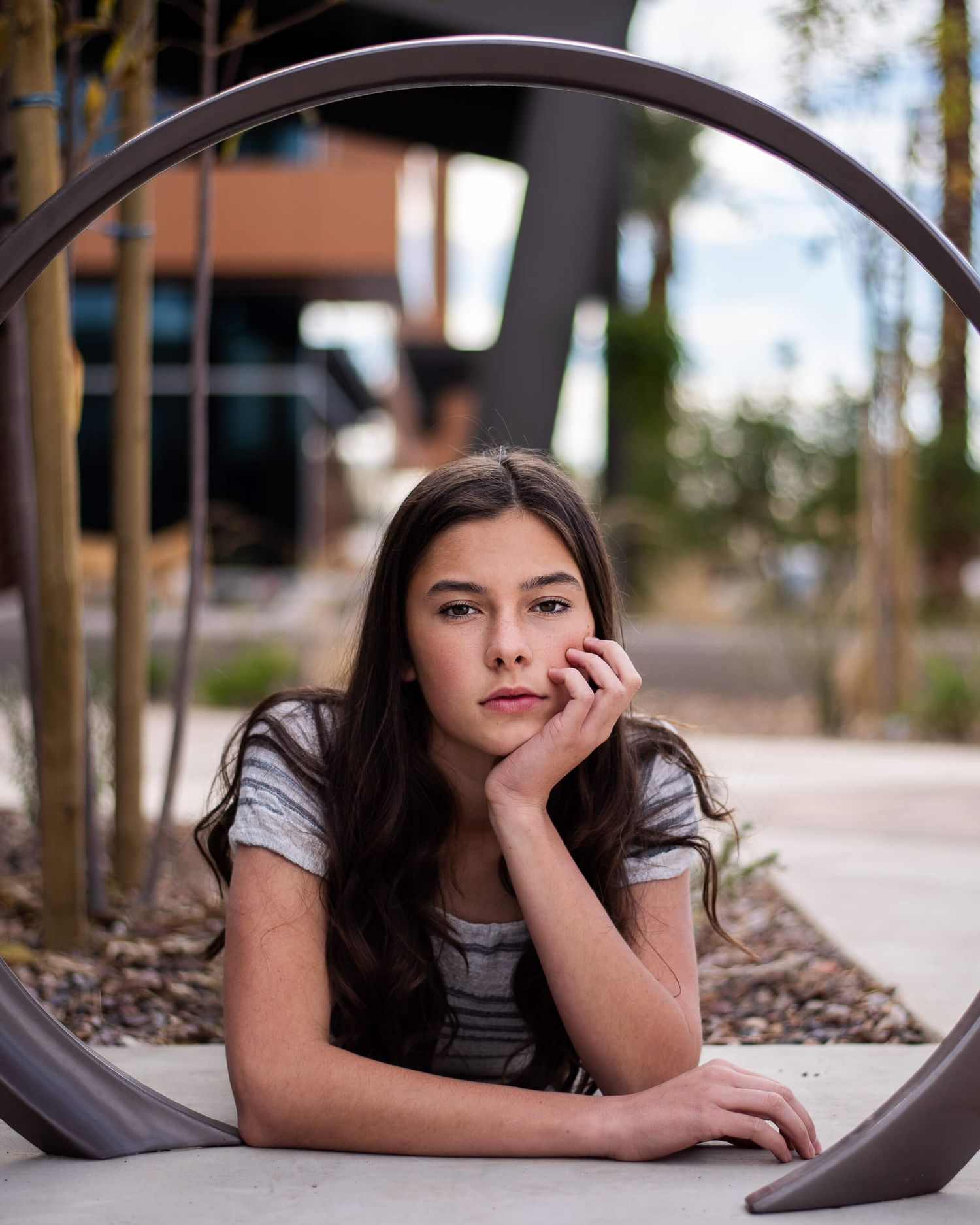 A young girl with long dark hair rests her chin on her hand, posing thoughtfully under a metal arch outdoors.