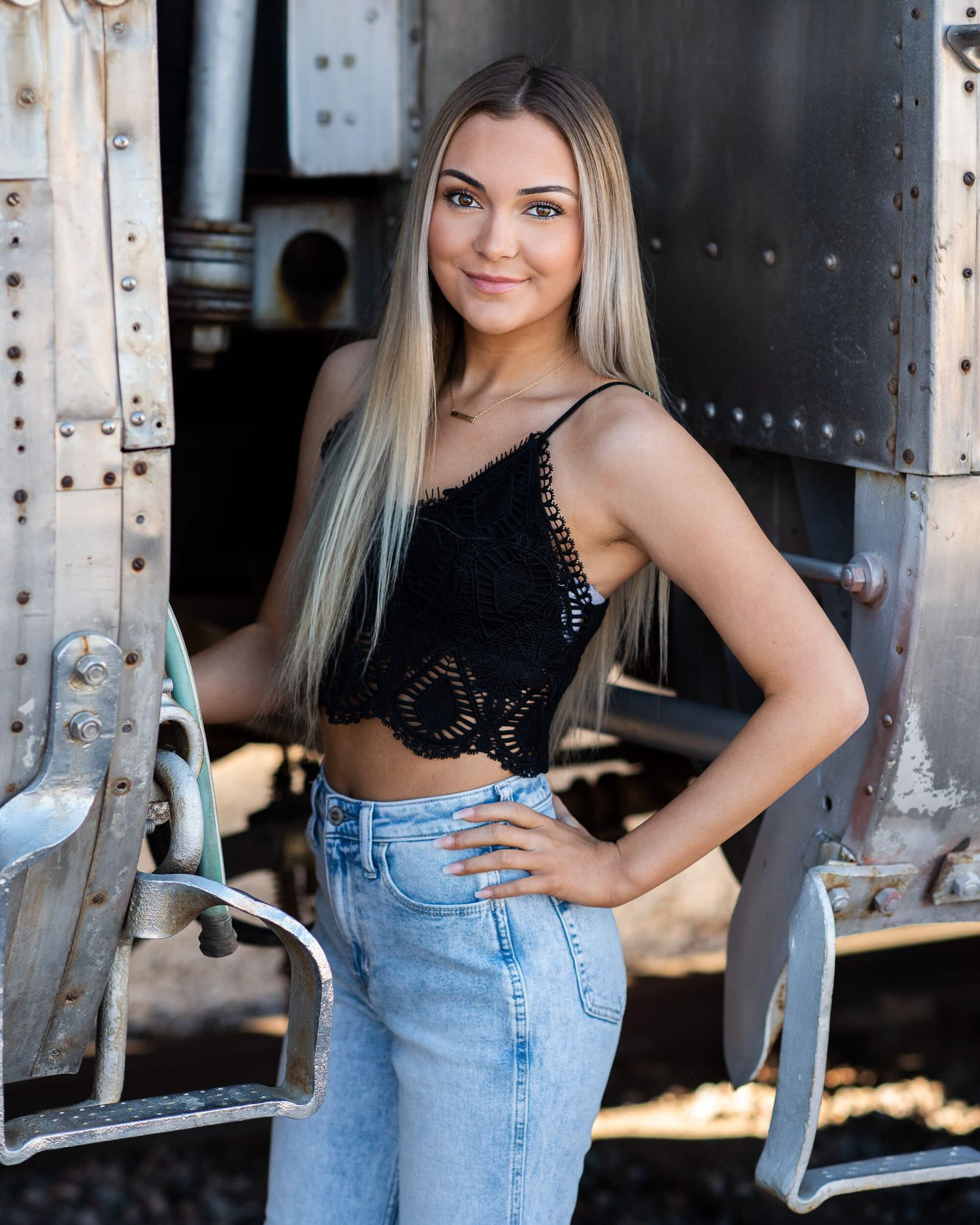 Blonde woman in black lace crop top and jeans posing confidently beside a vintage train car.