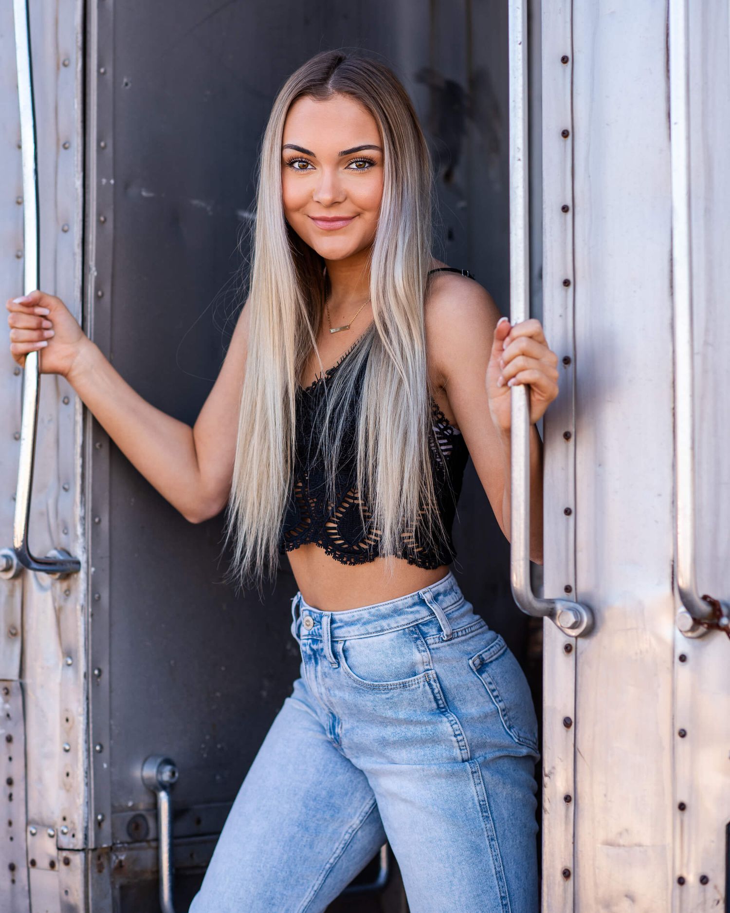 Young woman with long blonde hair poses in black crop top and jeans between train car doors.