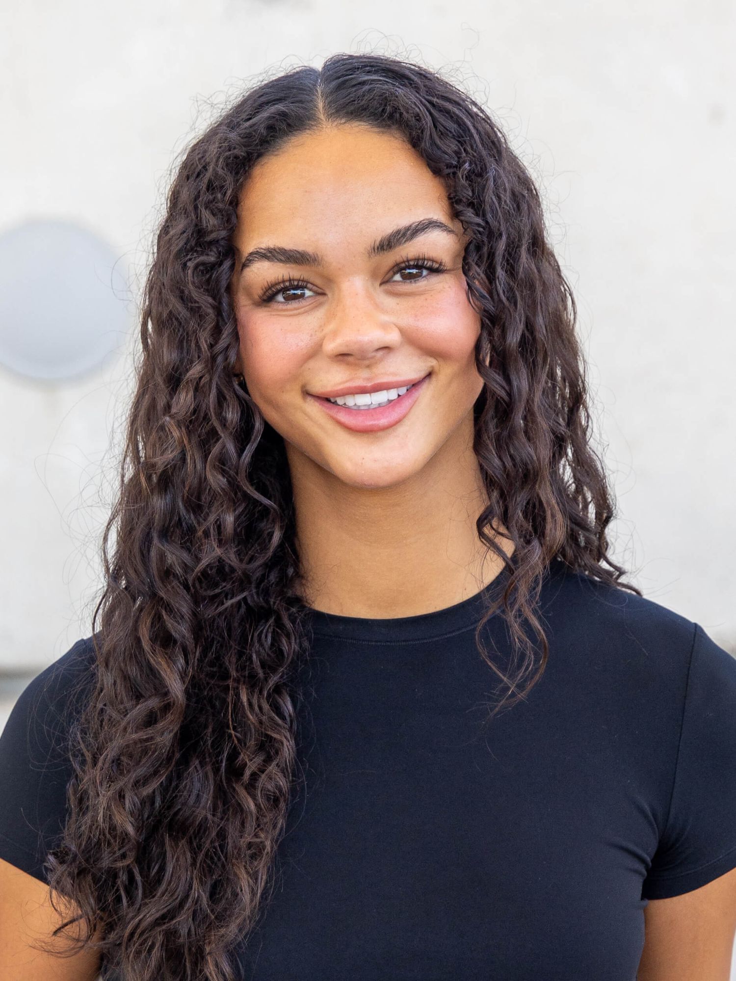 Young woman with long curly dark hair smiling confidently, wearing a black t-shirt against a neutral background.