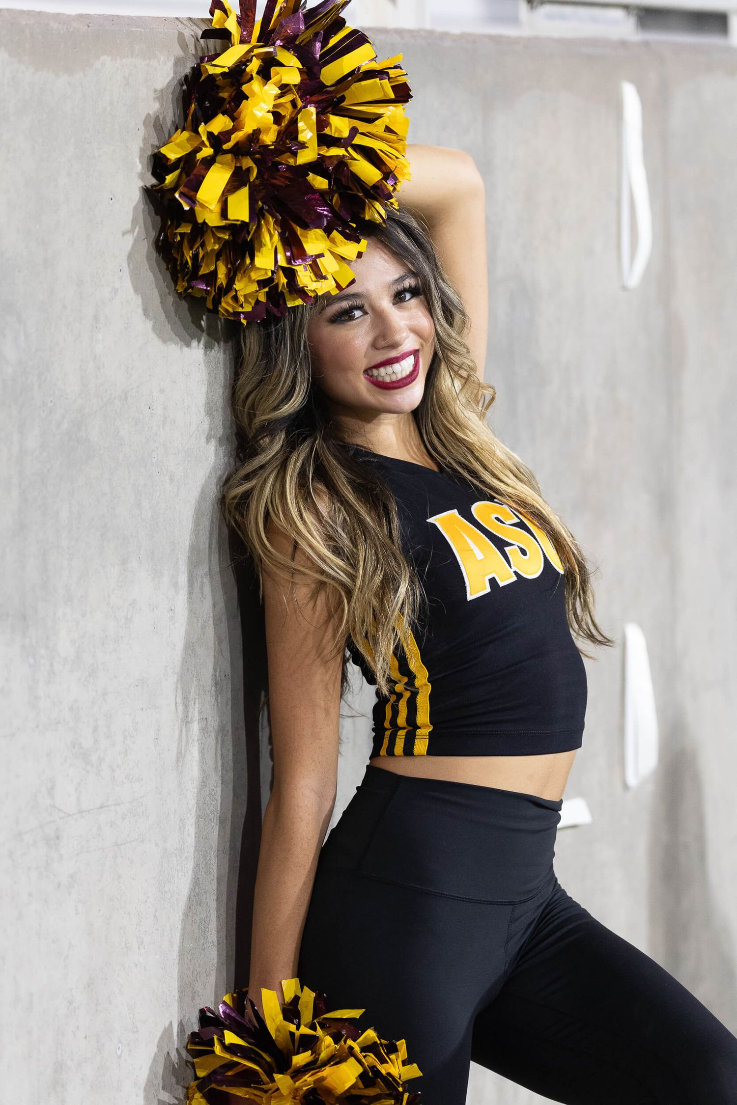 ASU cheerleader in black uniform poses with maroon and gold pom-poms against a concrete wall.