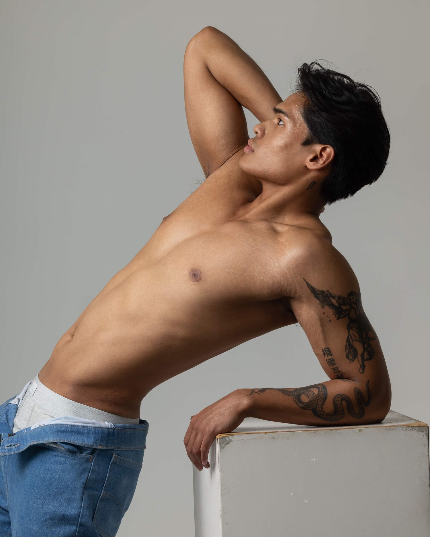 Lean tattooed man leaning back on white cube in studio setting.