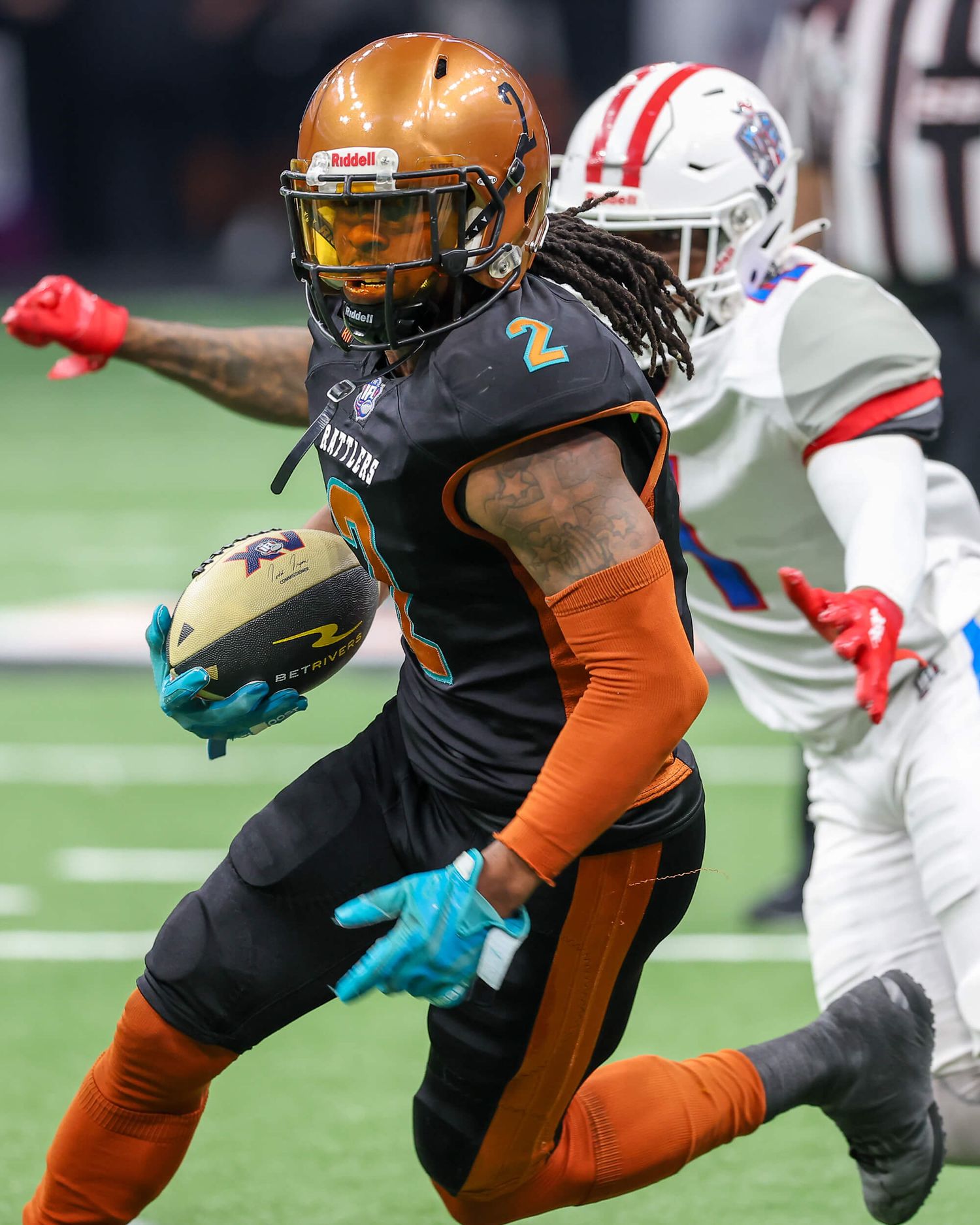 Player number 2 in orange uniform runs with football during indoor arena football game.