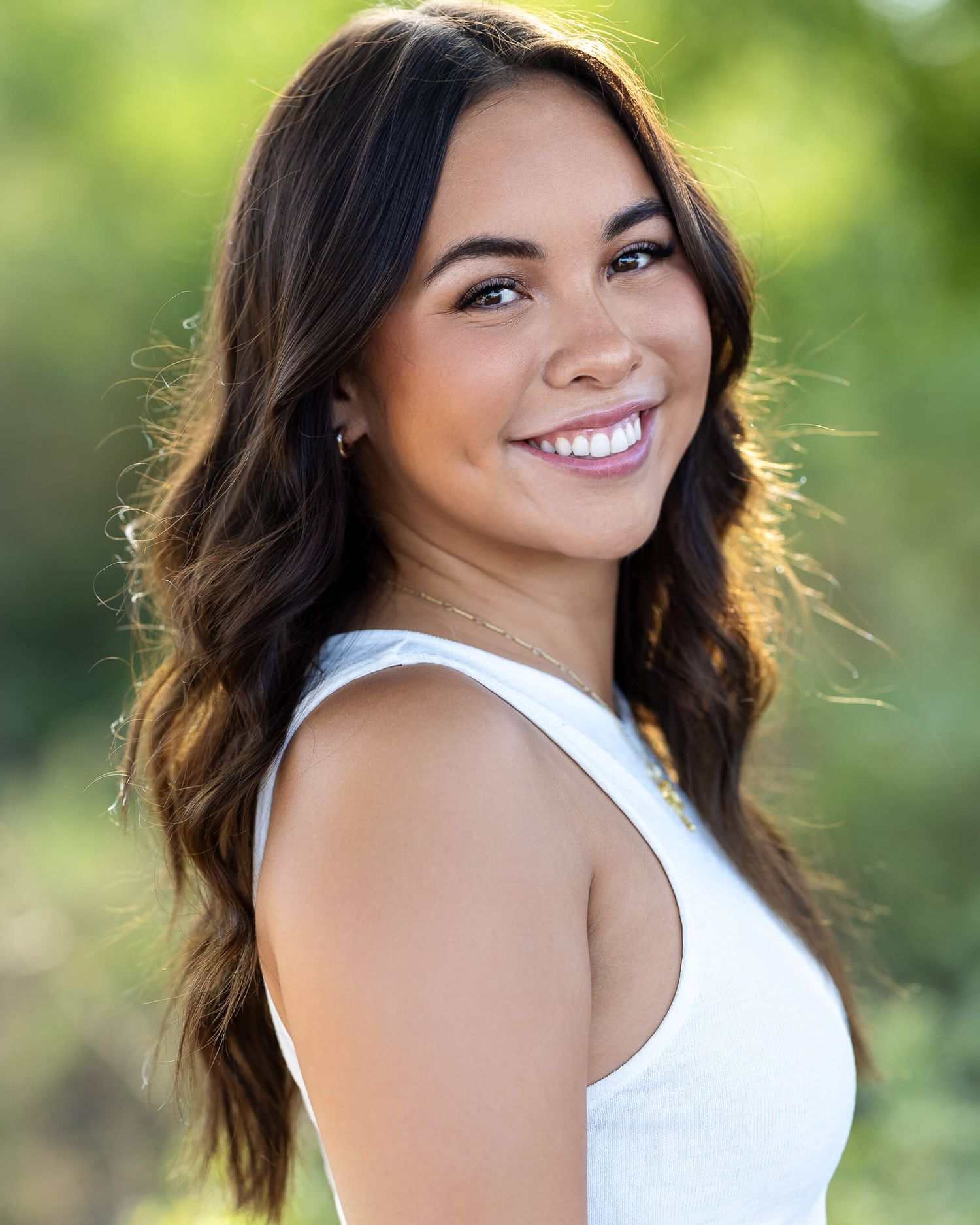 Smiling young woman with wavy brown hair poses outdoors in a white top.