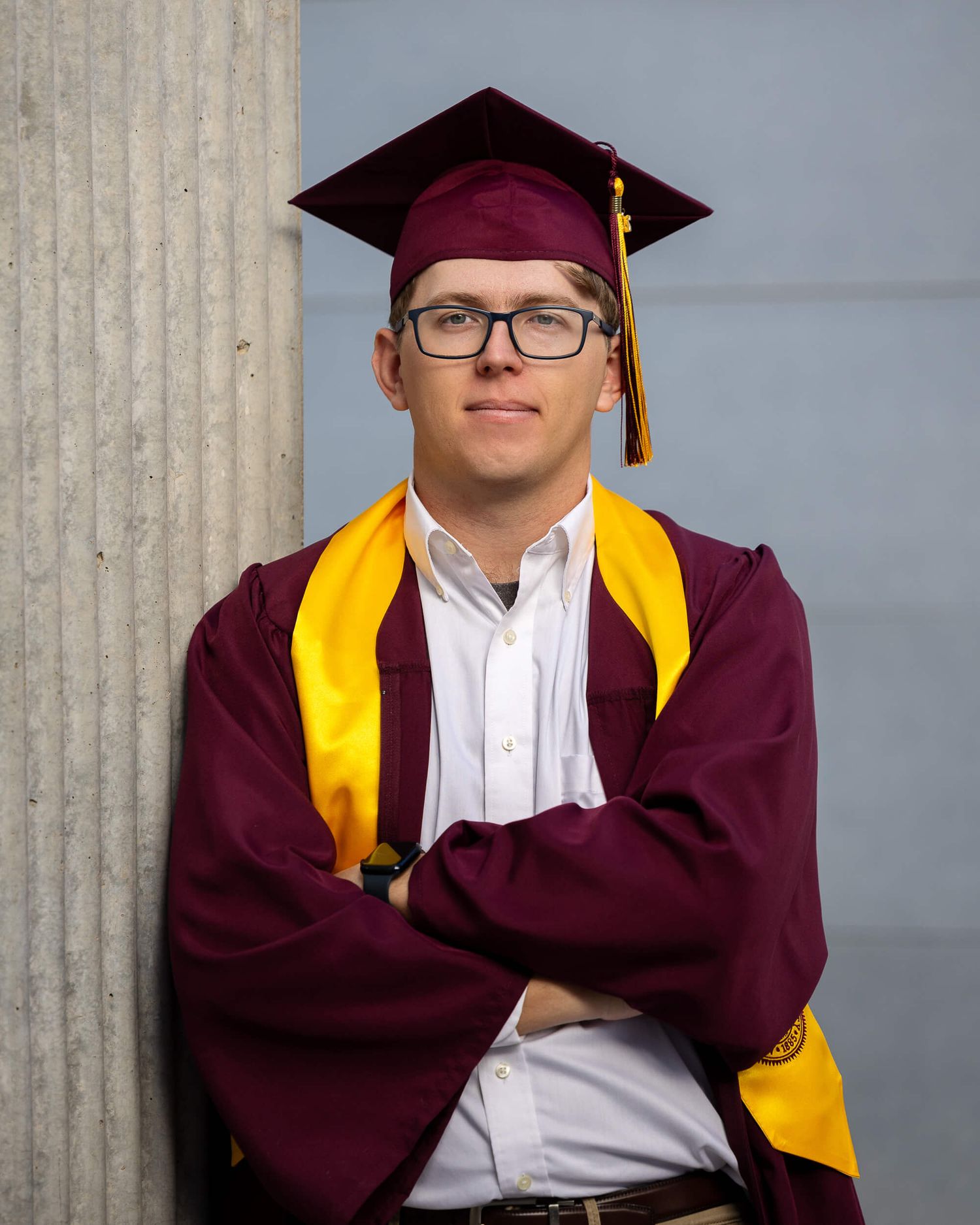 Male graduate in maroon ASU cap and gown with gold stole leaning against column.