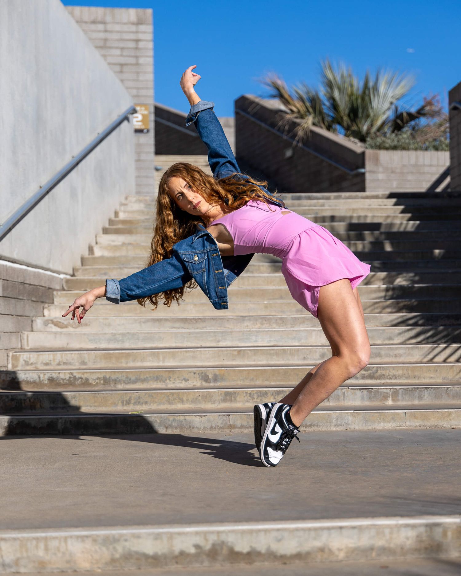 Young dancer in pink skirt and denim jacket leaping on outdoor stairs.