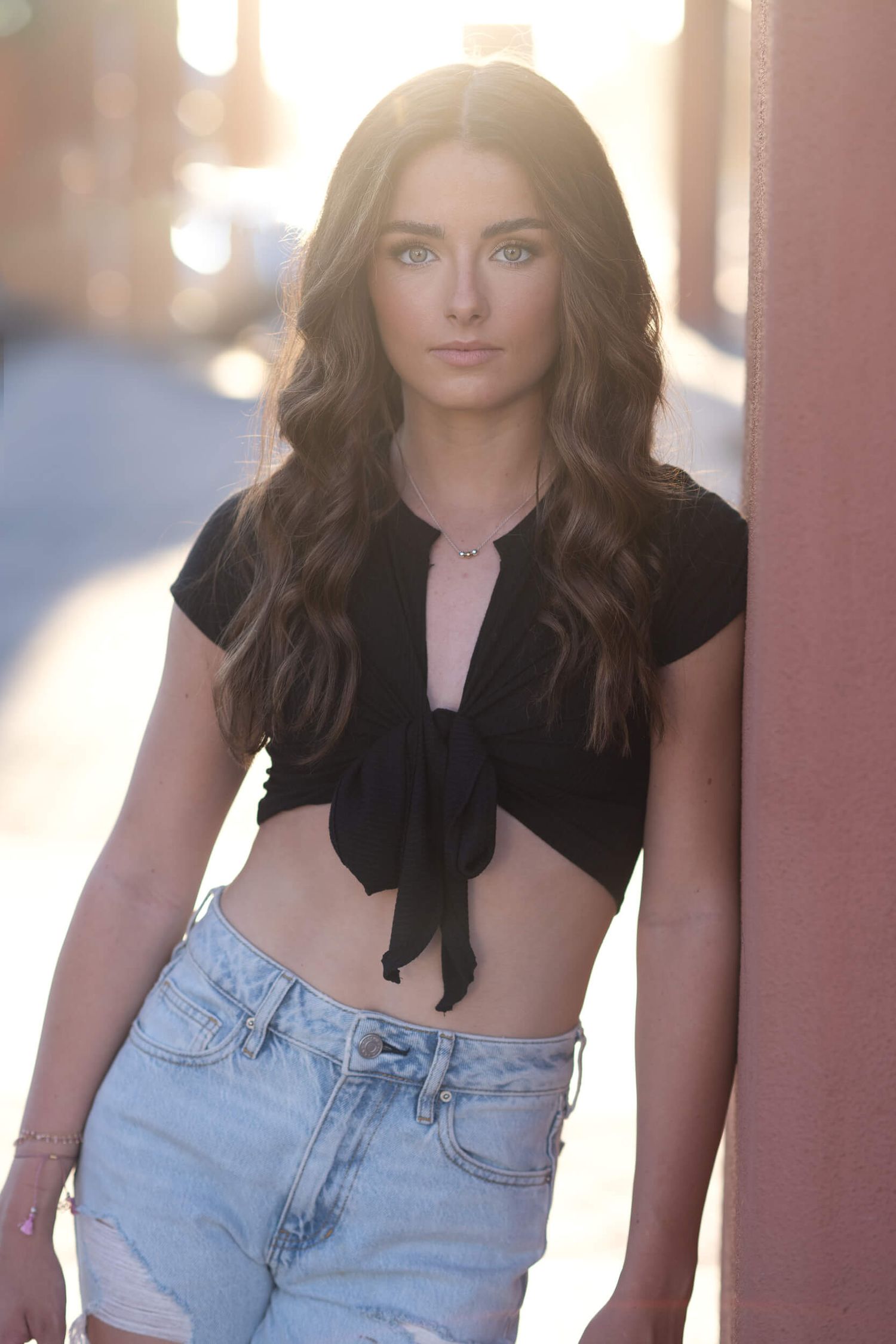 Brunette woman in black tied crop top and denim shorts posing in an alley at sunset.
