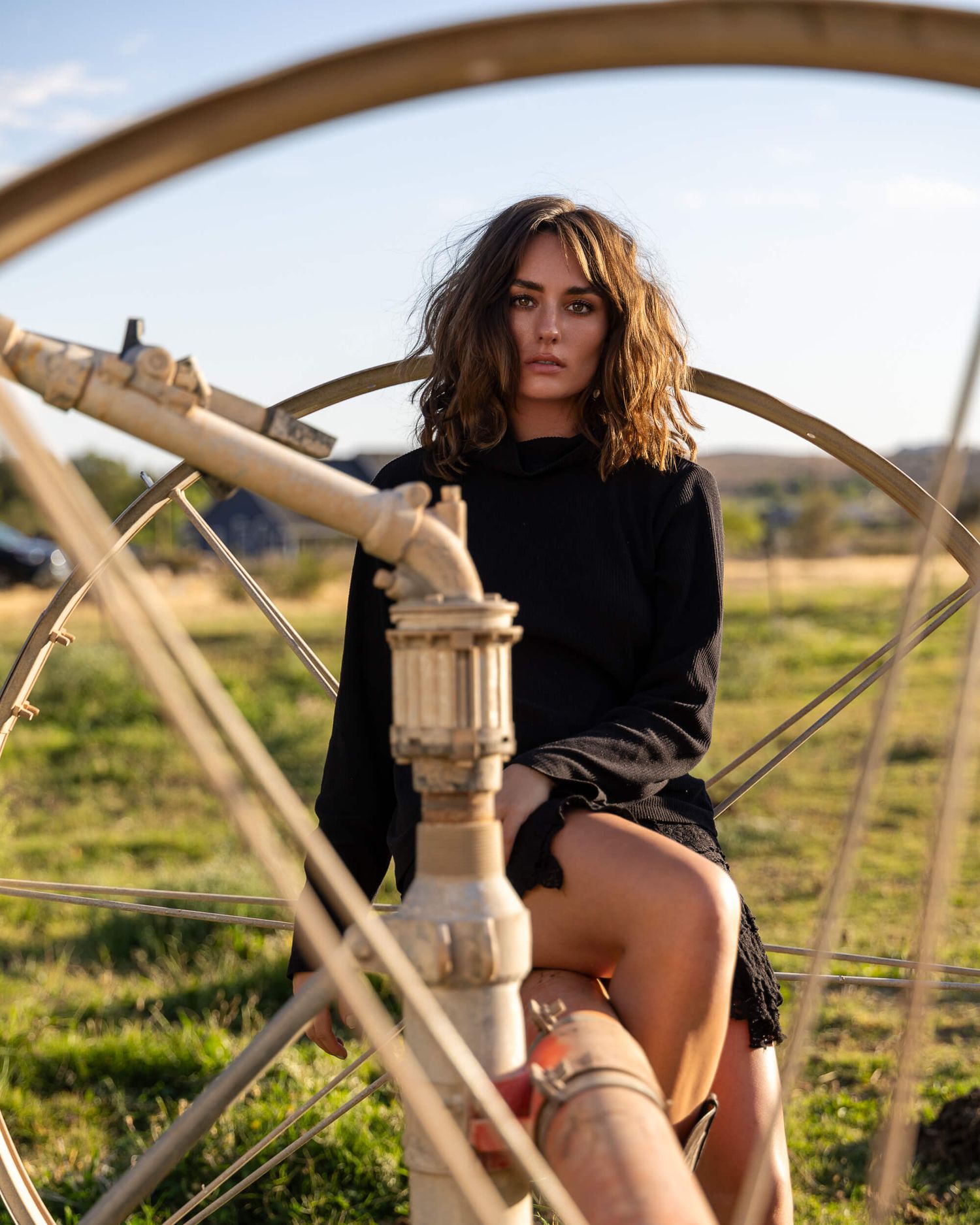 Young woman in black sweater and mini skirt sitting on vintage farm equipment in a field.