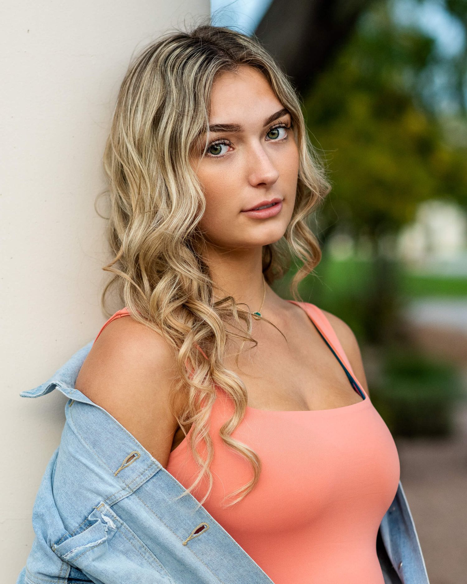 Young blonde woman in peach top and denim jacket leaning against a column outdoors.